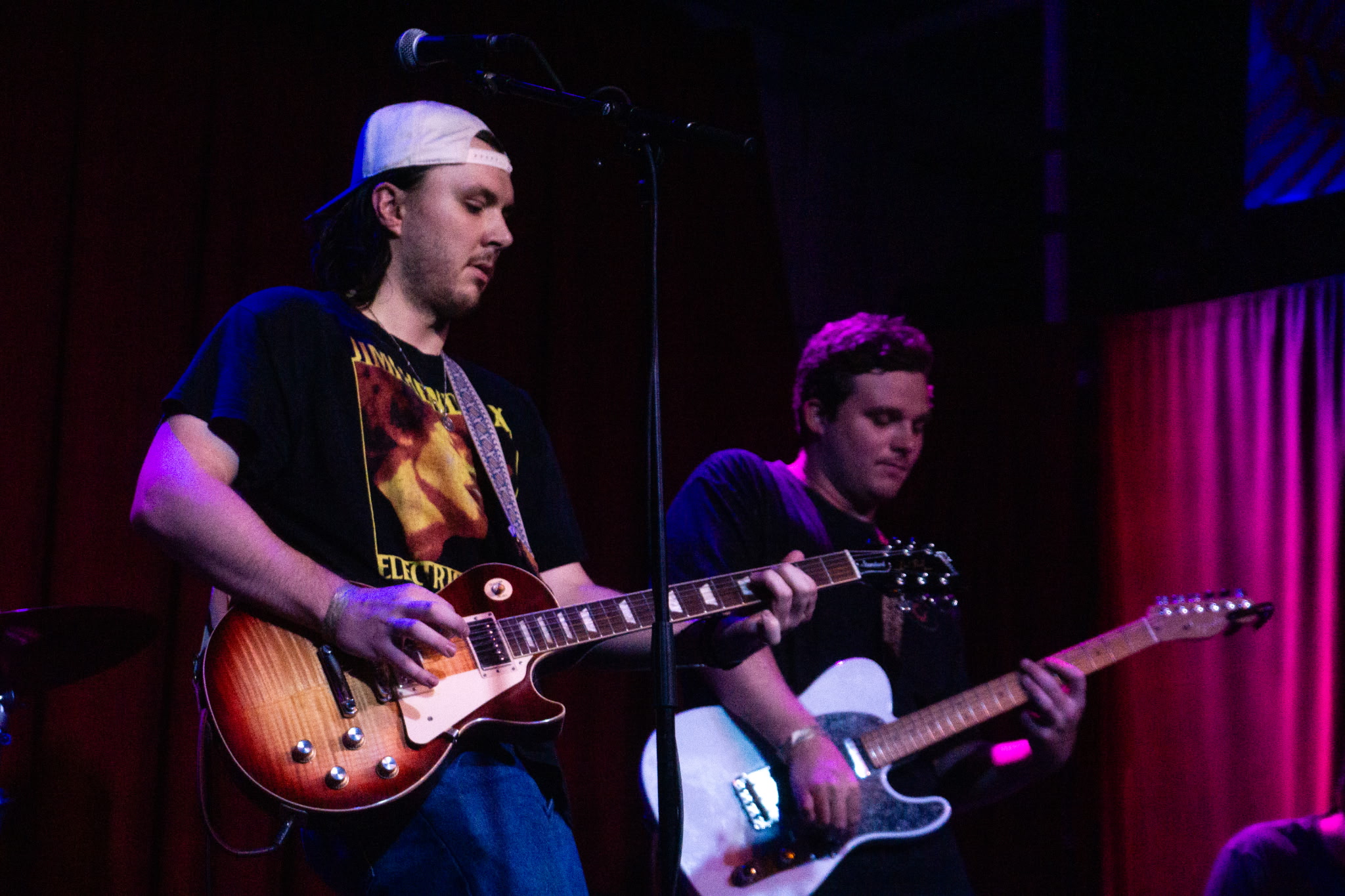 Two young men playing electric guitars on stage with red curtains and purple lighting in the background.