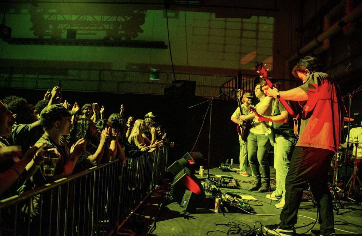 Musicians perform on stage with audience watching, some raising hands, in a concert setting with green lighting.