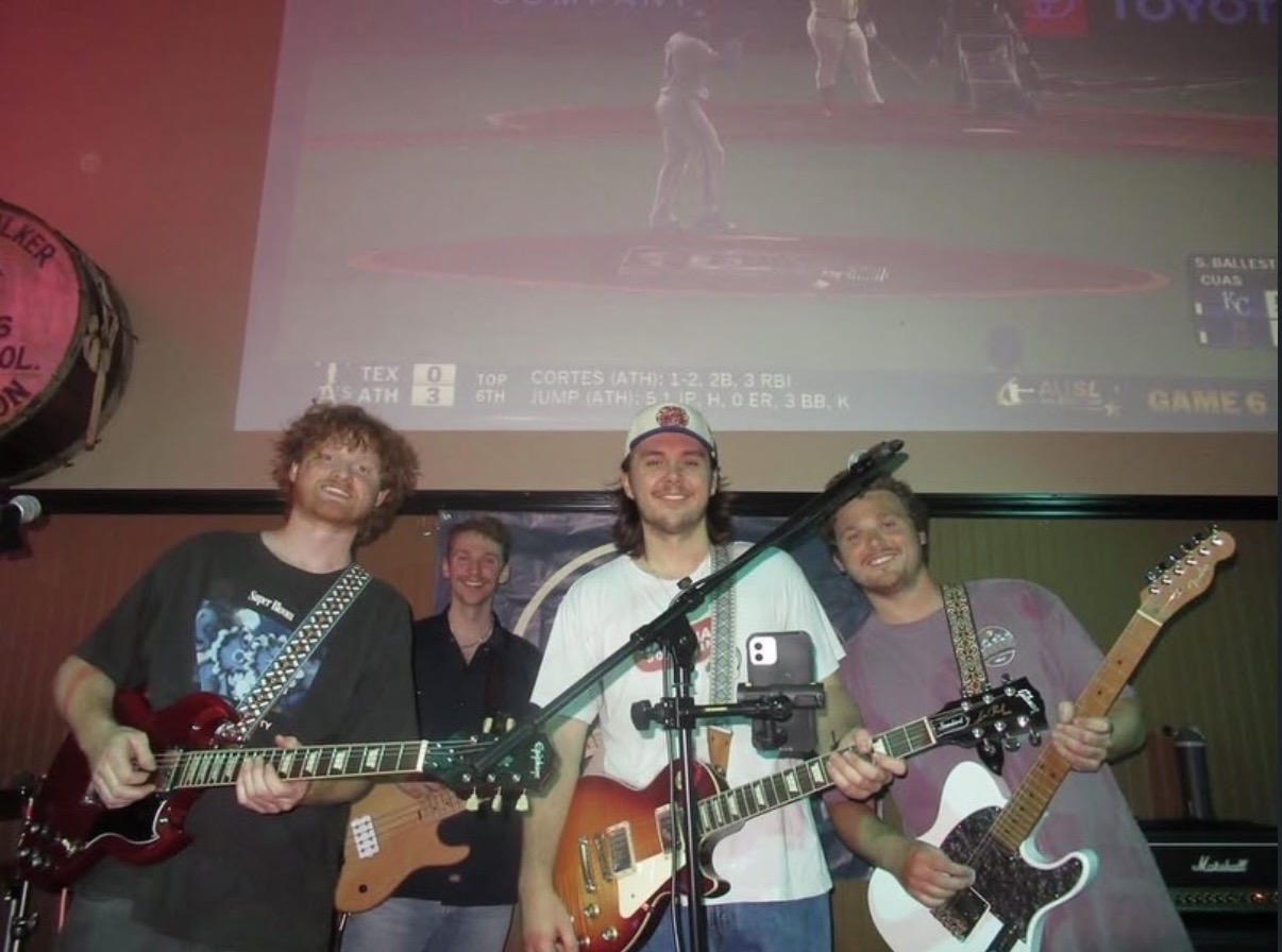 Four people playing guitars on stage with a large screen behind them showing a baseball game.