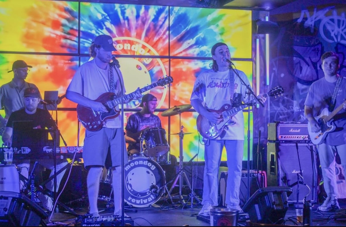 Band performing on stage with colorful background, five musicians playing guitars, drums, and keyboard, stage lights visible.