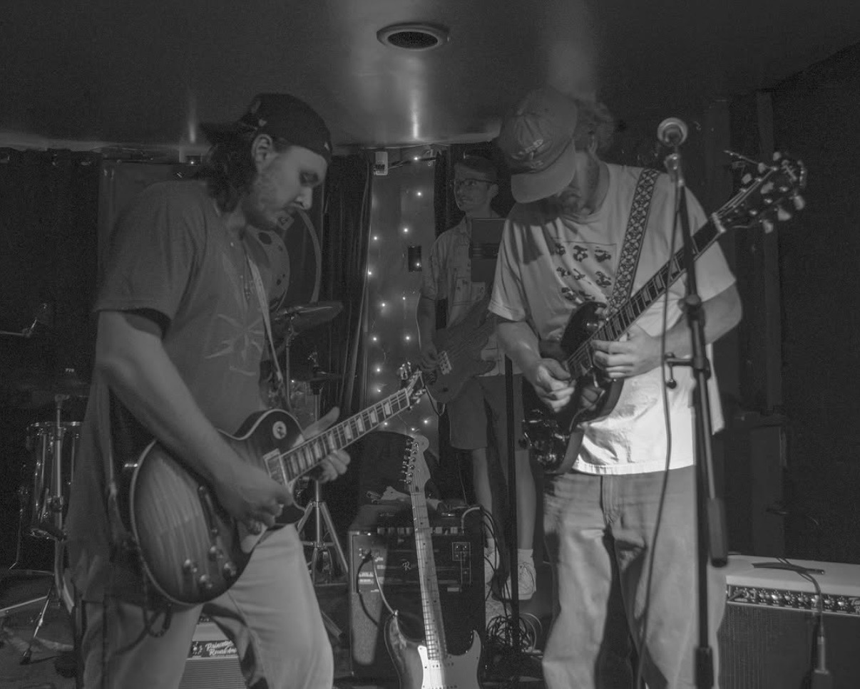 Three musicians perform on stage with guitars, one wearing a cap, in a dimly lit setting.