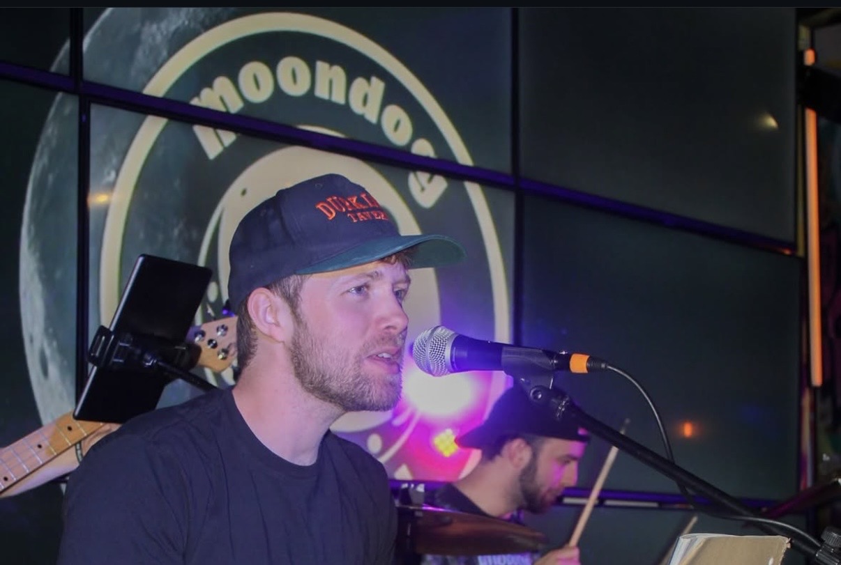 Man with beard wearing a cap singing into a microphone, with a person playing drums in the background, in a music venue.