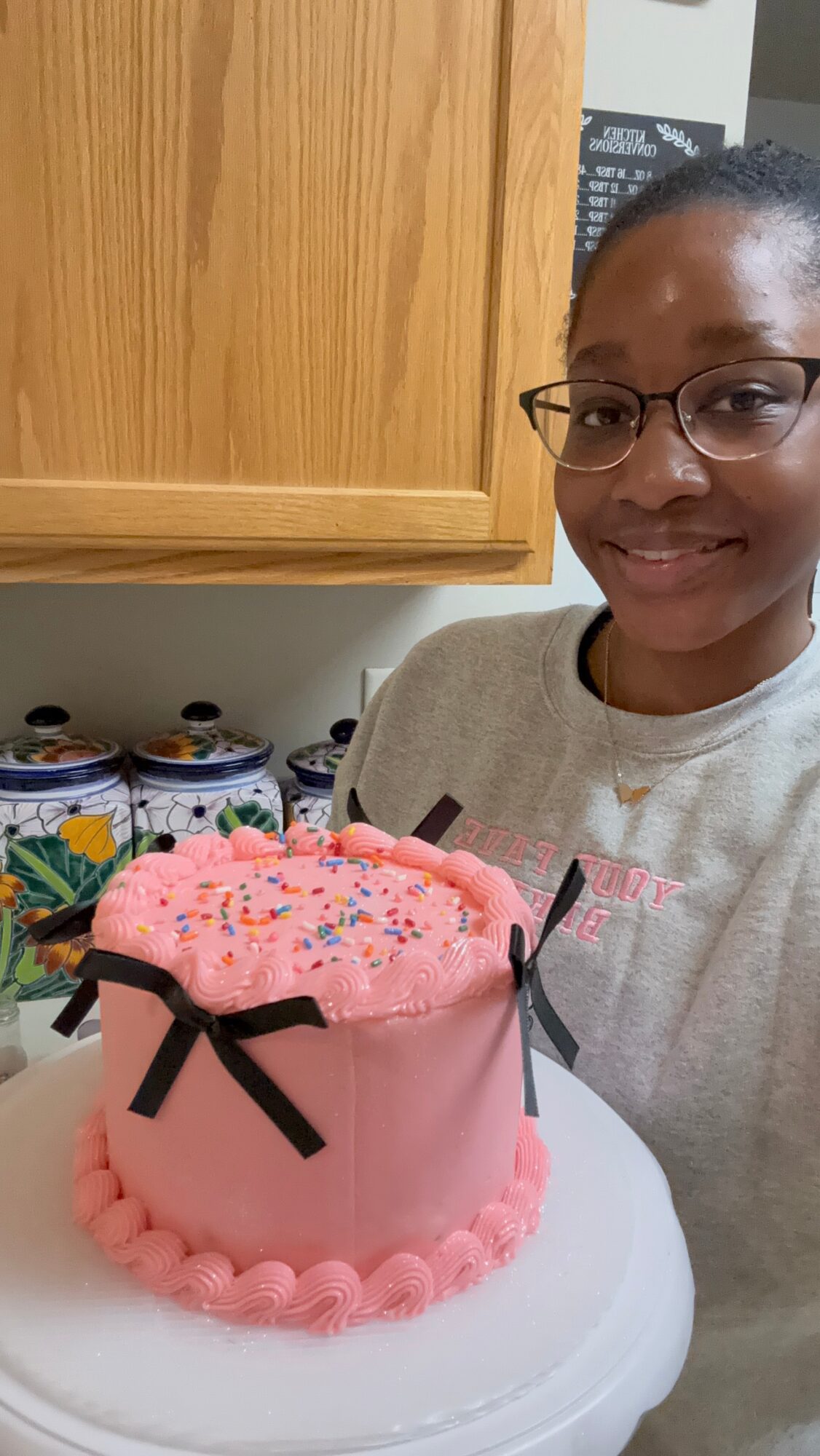Smiling woman with glasses stands behind a pink birthday cake with sprinkles and black bow decorations.