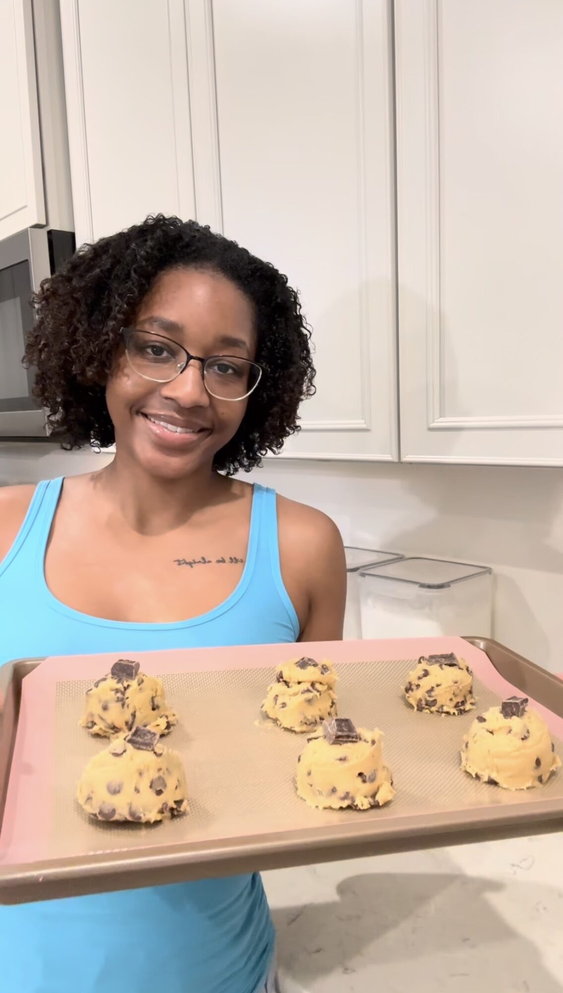 Young woman with glasses and curly hair smiling in a kitchen, holding a tray of six cookies with yellow and chocolate chips.