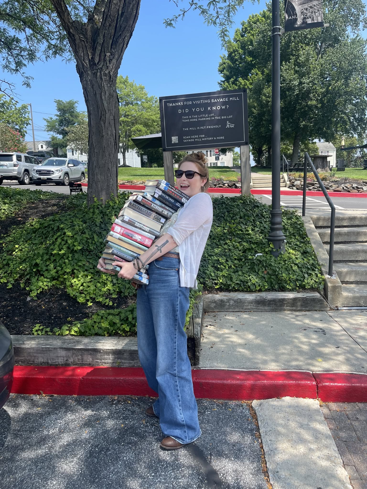 Person holding multiple stacked books standing outdoors near a tree and a sign, with cars in the background.