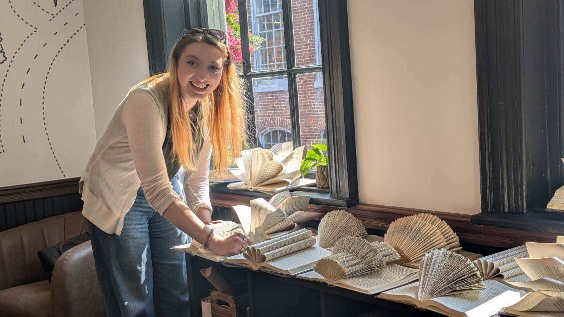 Person with long hair smiling, standing by a window with open books and paper fans on a table.