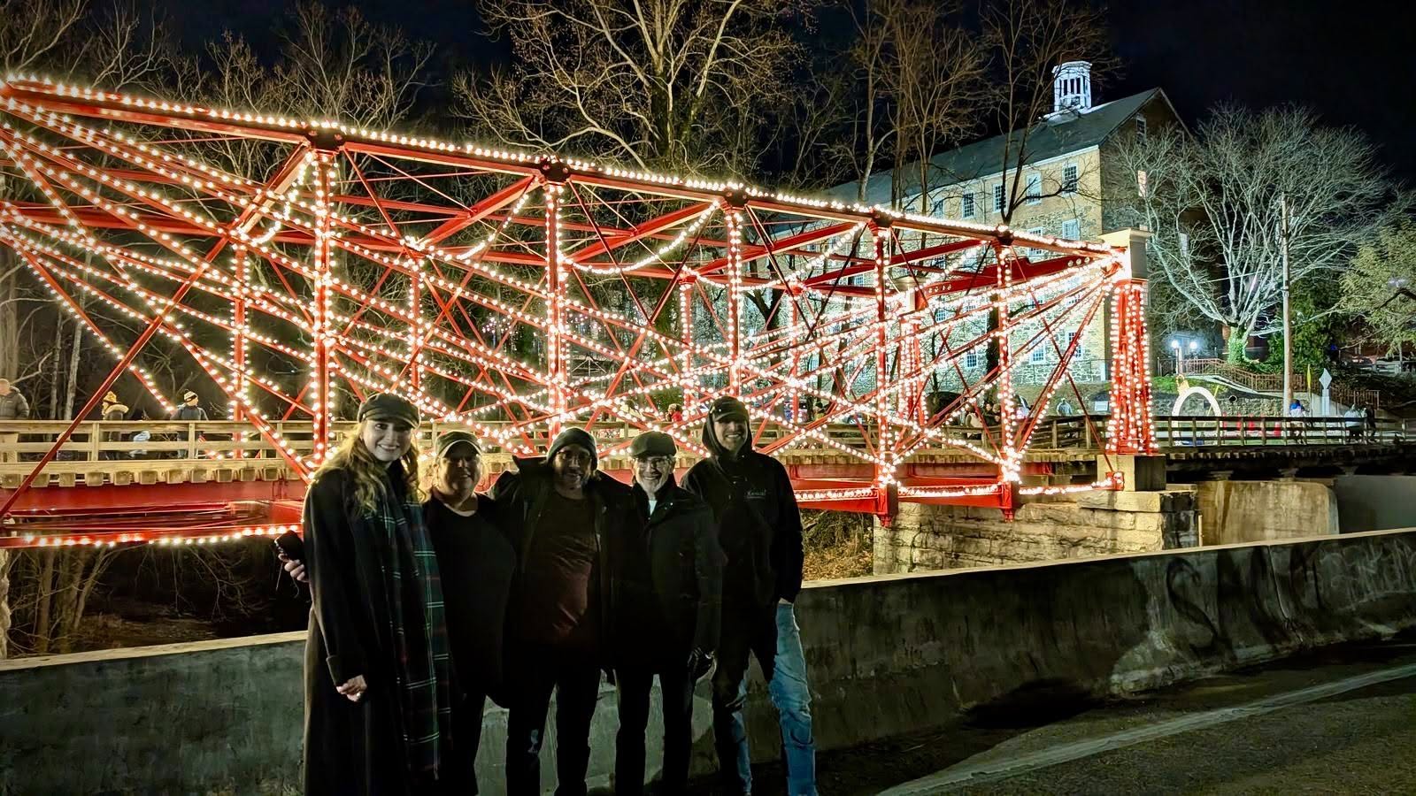 Group of five people standing near a lit amusement ride at night, with trees and a building in the background.