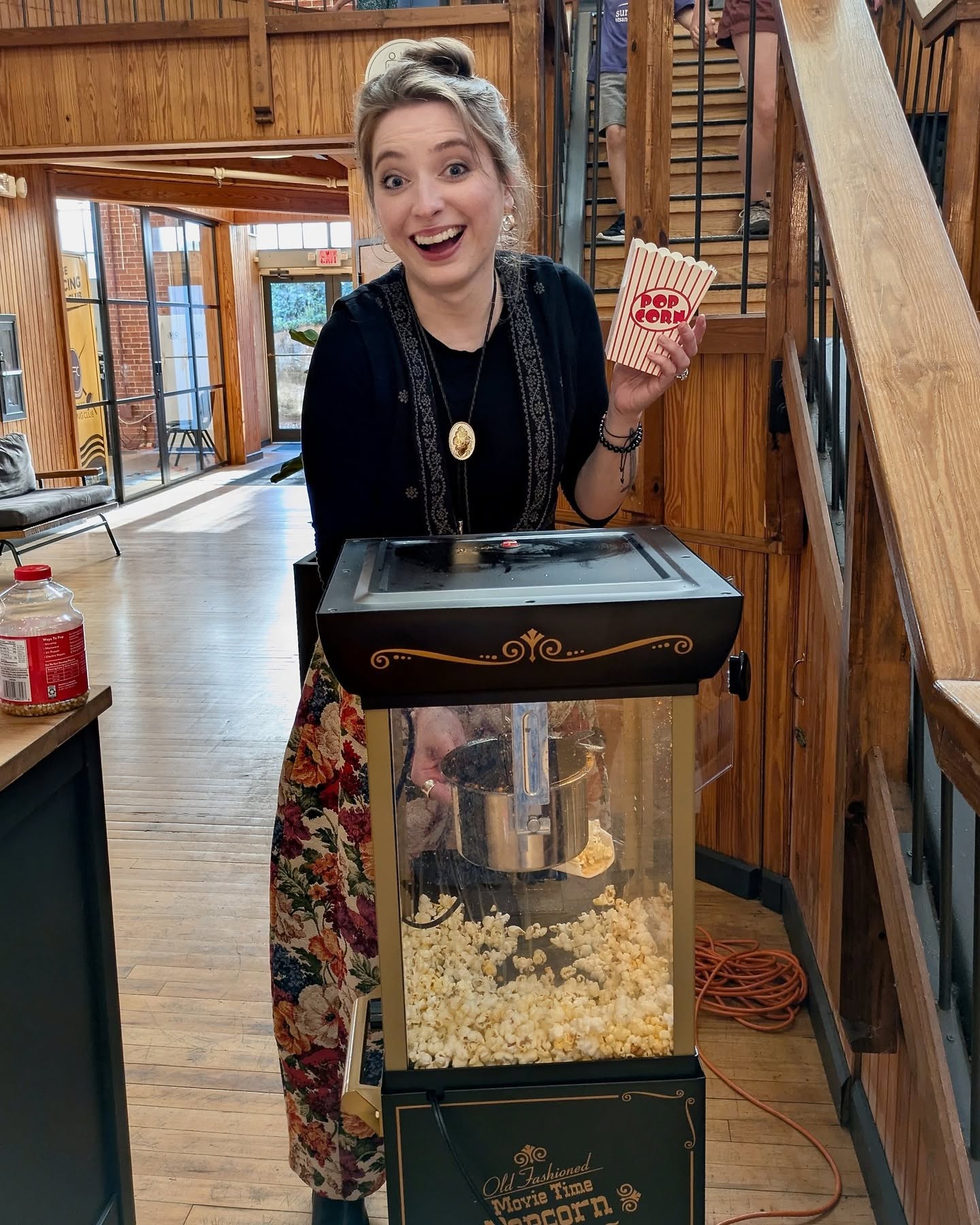 Young woman with a bun hairstyle holding popcorn and smiling indoors, wooden interior, staircase in background.