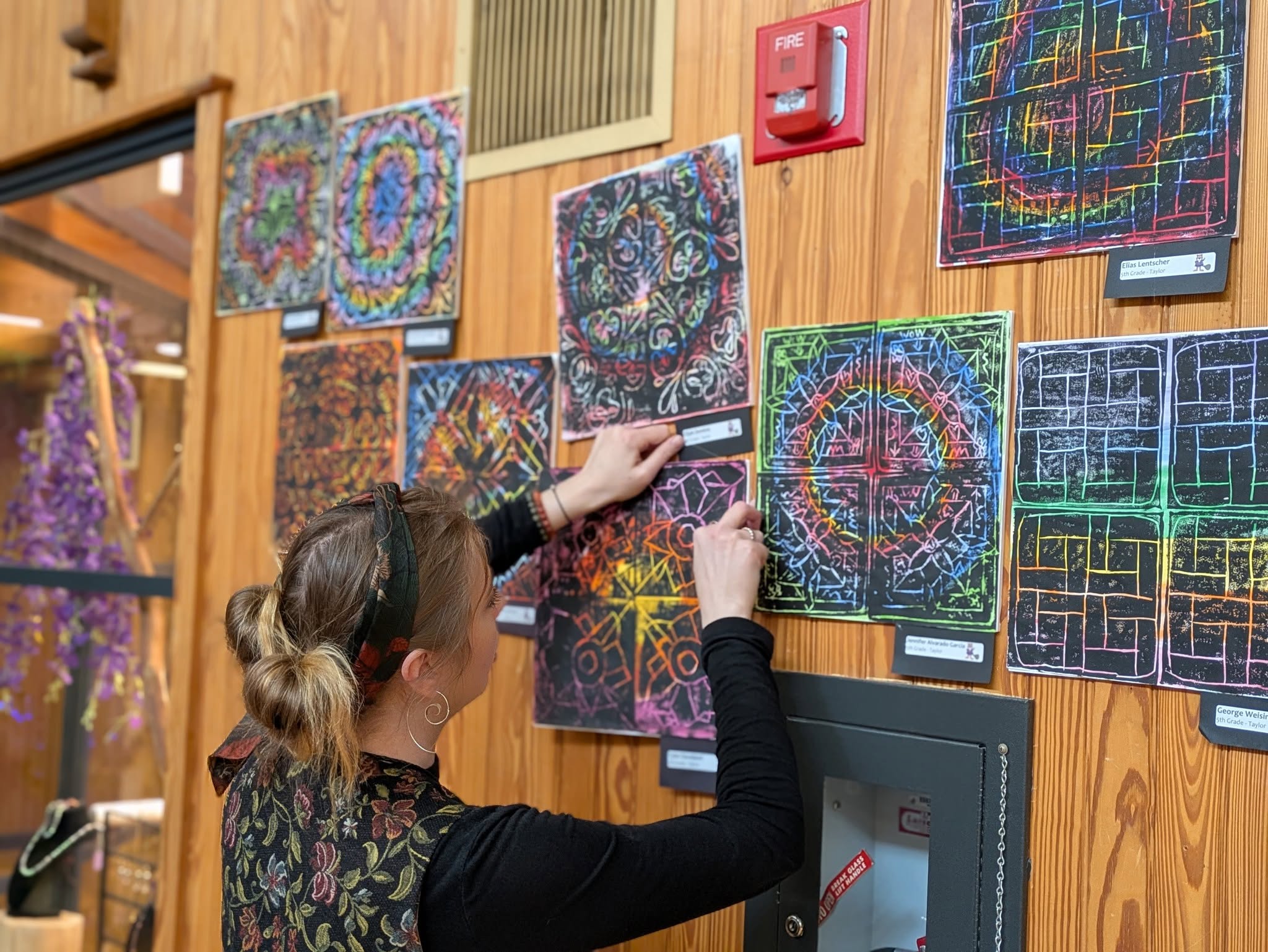Person with blonde hair in a bun, wearing a black and floral top, hangs colorful abstract artwork on a wooden wall.
