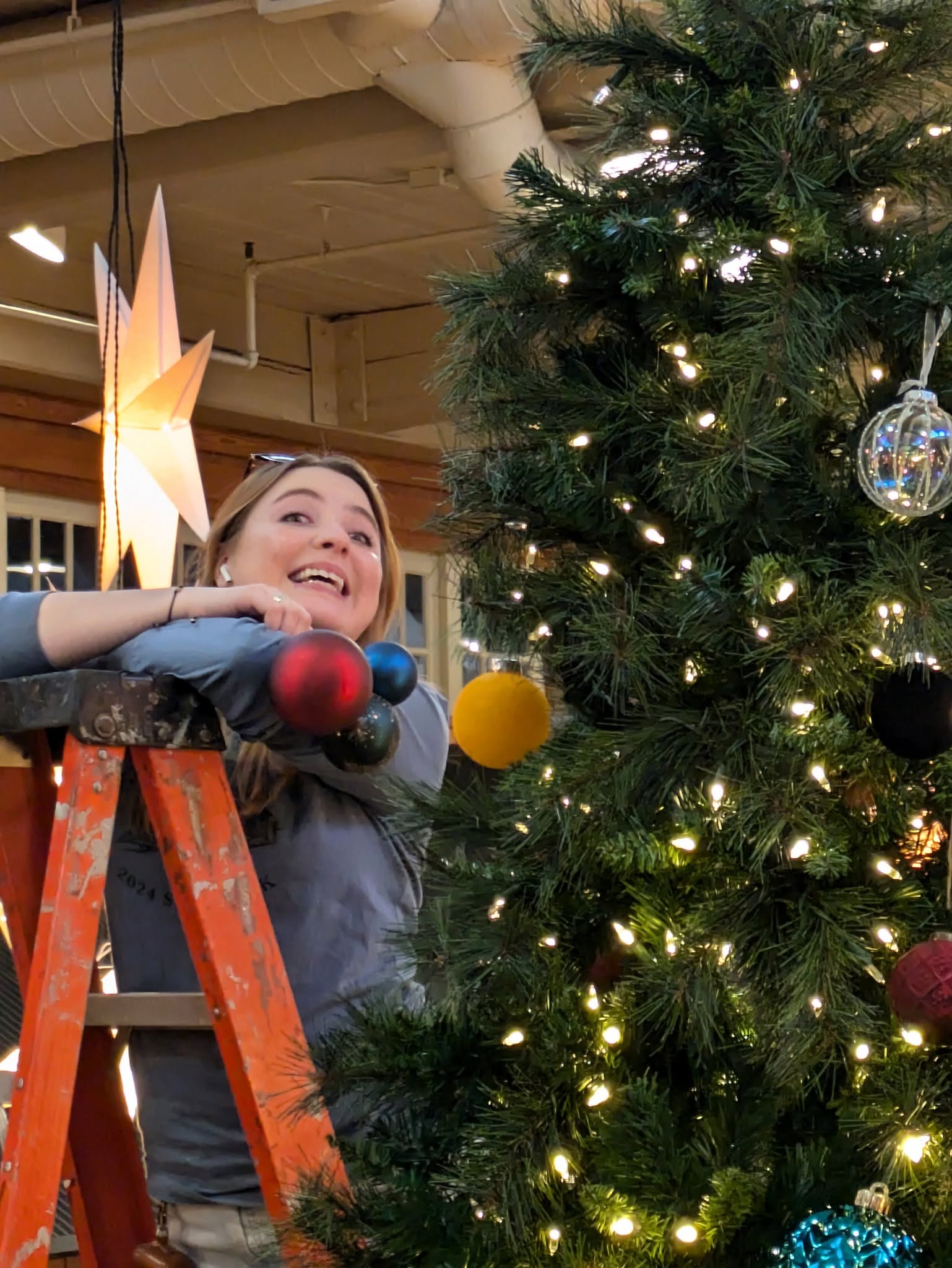 Woman decorating a Christmas tree with ornaments, smiling, indoors with festive lighting and decorations.