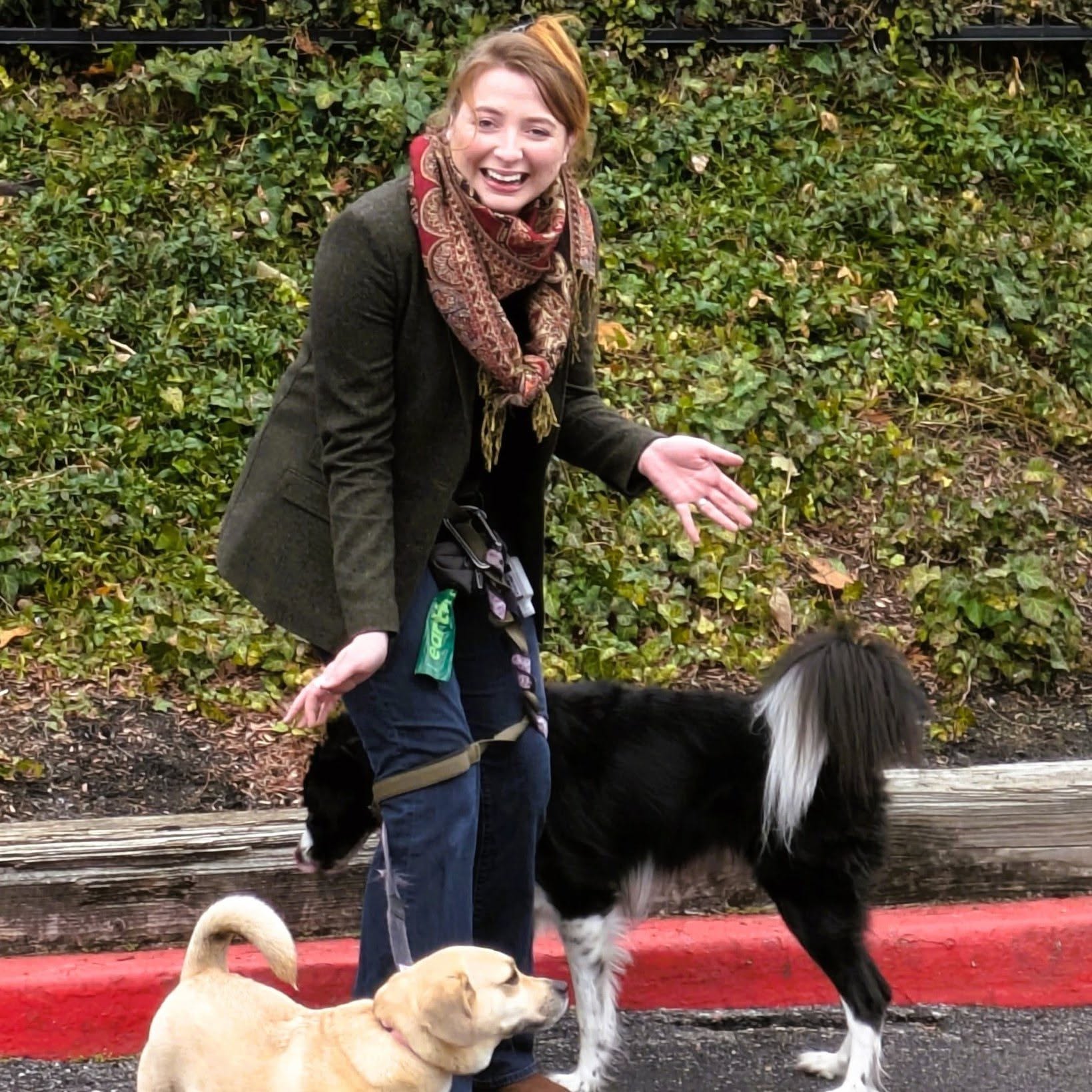 Young woman smiling outdoors with a black and white dog and a light-colored dog, standing on a sidewalk with greenery behind.