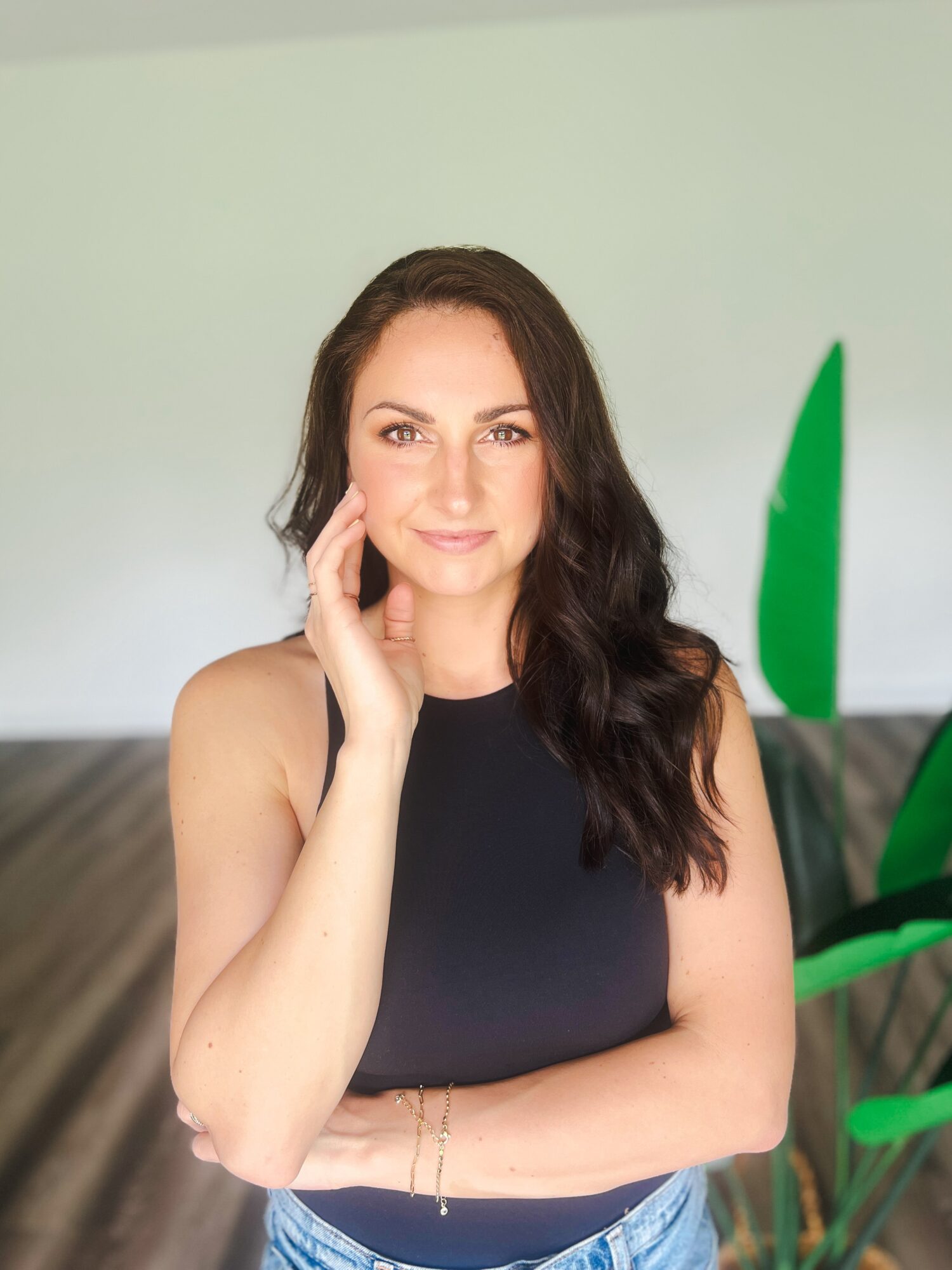 Woman with long dark hair wearing a sleeveless black top, smiling, standing indoors with green plants in background.