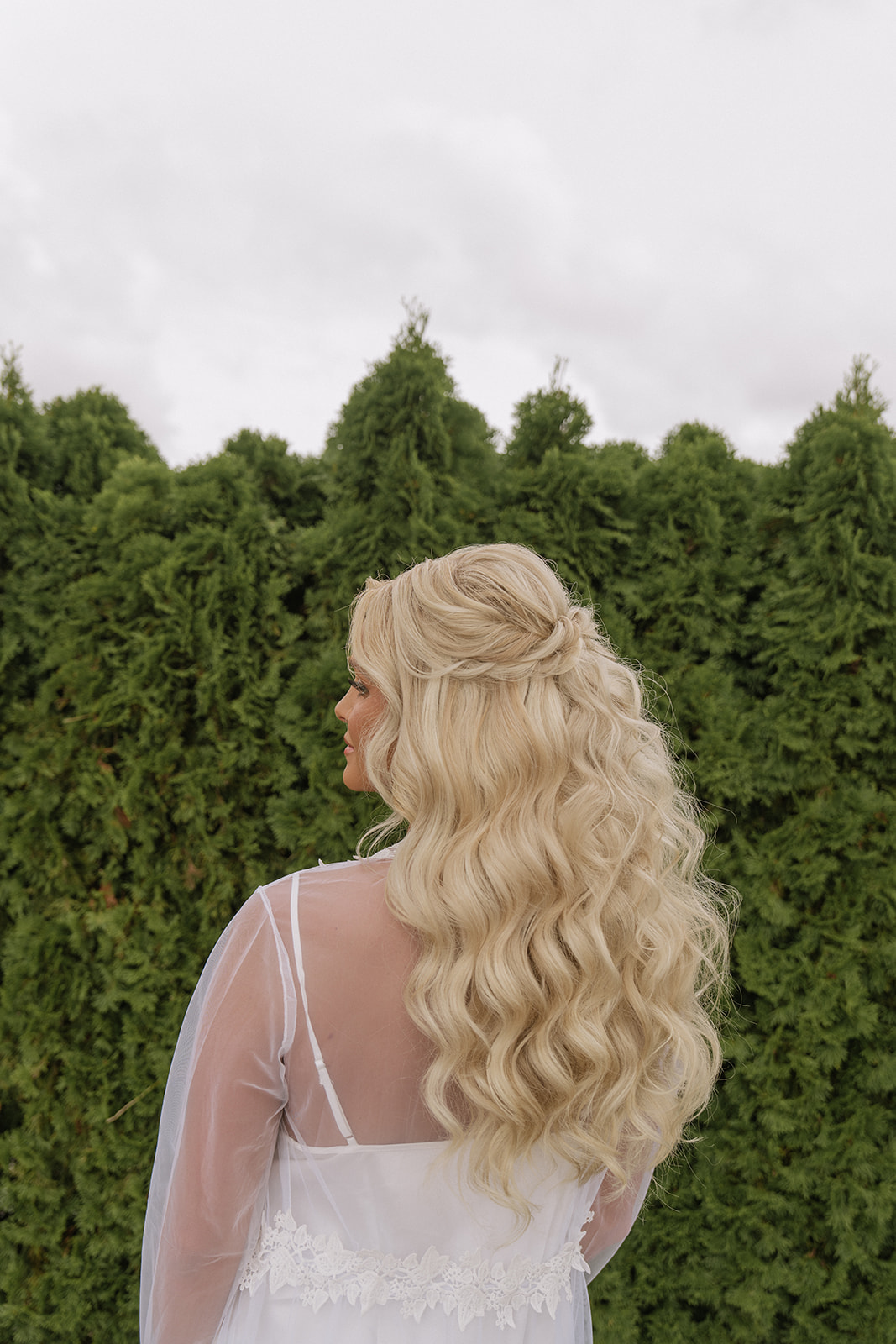 Person with long, wavy blonde hair standing outdoors with green trees in the background, facing away from the camera.