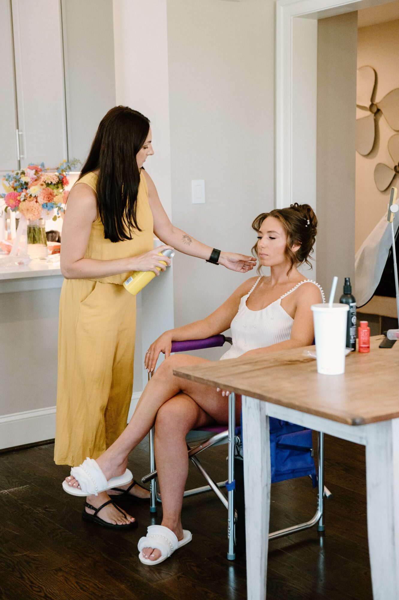 Two women in a room, one sitting in a chair and the other standing, with flowers and makeup items nearby.