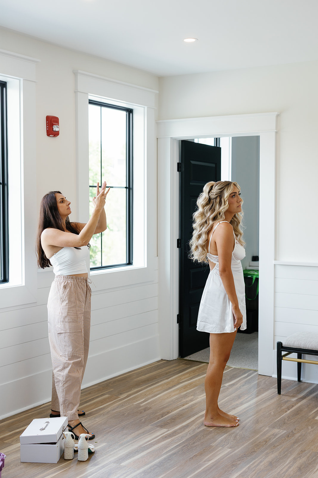 Two women in a bright room, one taking a photo or video with a phone, the other standing barefoot, facing a mirror.