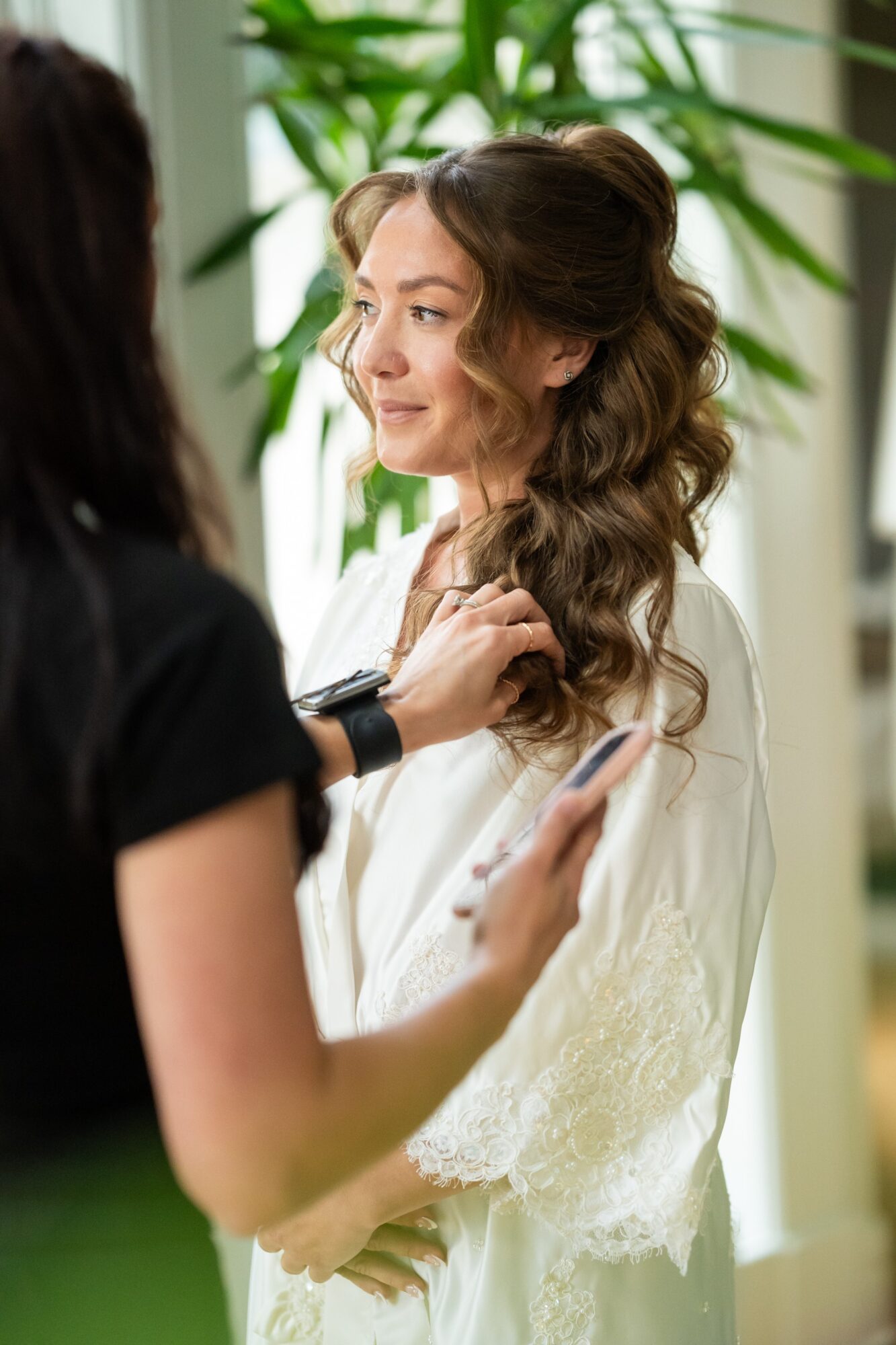 Woman with long, wavy hair smiling as another person adjusts her hair, holding a smartphone, in a bright room with plants.