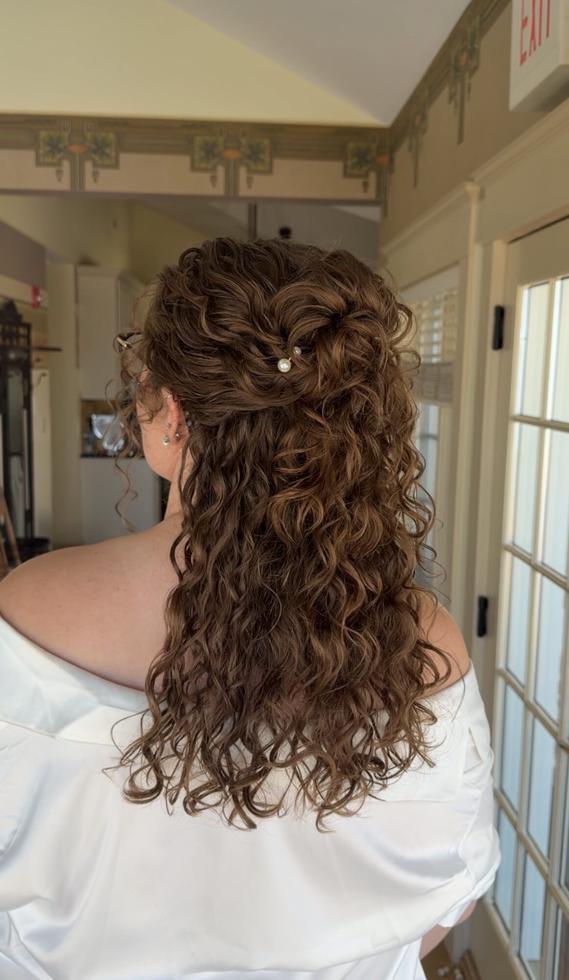 Back view of a woman with long, curly hair decorated with small white flowers, wearing a white top.