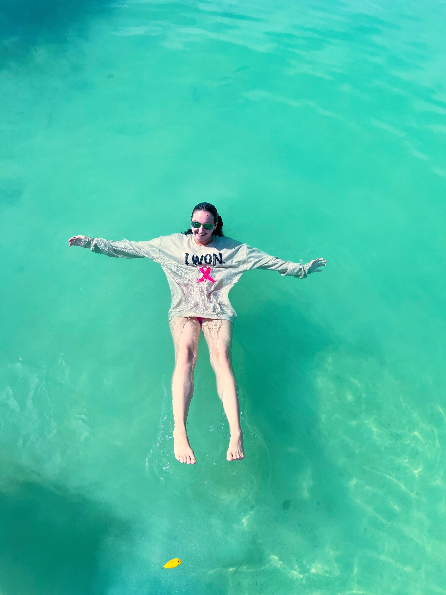 Person floating on water with arms outstretched, wearing sunglasses and a sweatshirt, in a turquoise body of water.