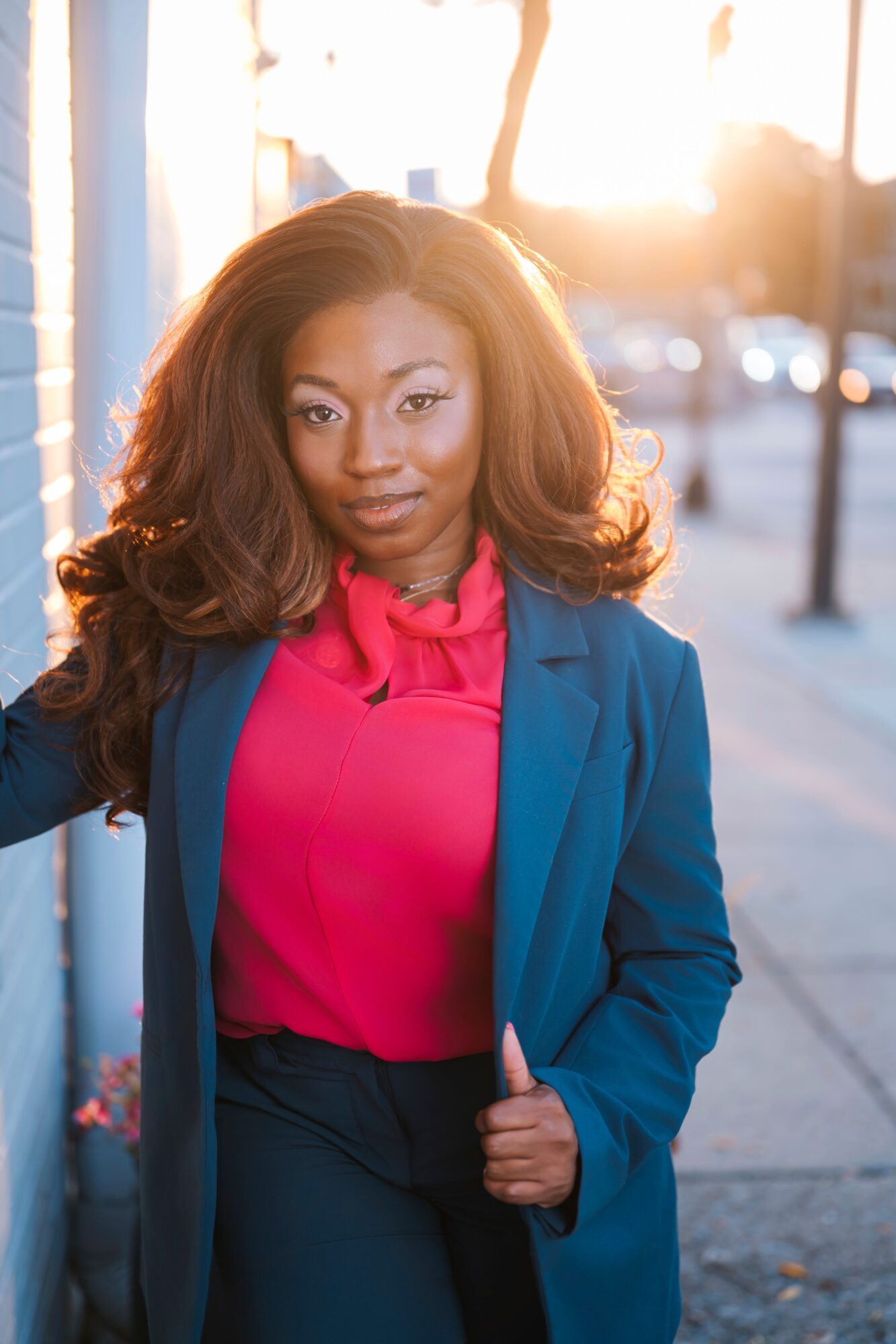 Woman with long hair wearing a pink blouse and blue blazer standing outdoors at sunset.