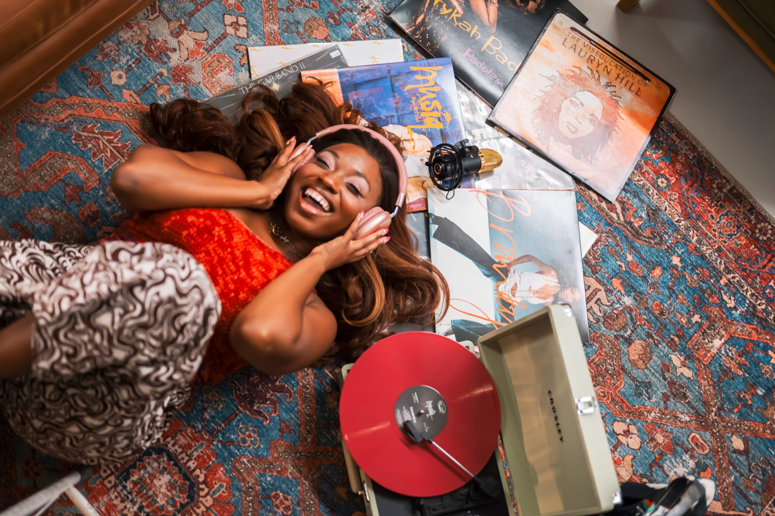 Woman lying on colorful rug, smiling, surrounded by vinyl records and music equipment, holding headphones to her ears.
