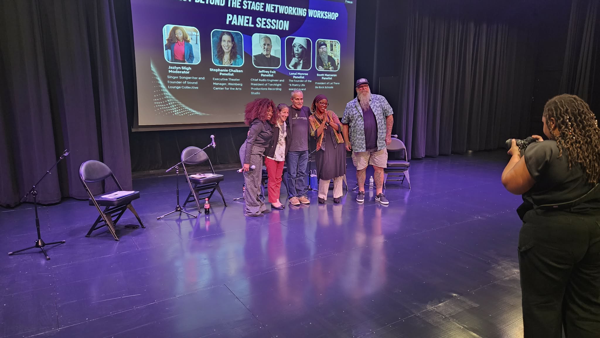 Five people stand on a stage in front of a large screen, with chairs nearby, and a woman taking a photo.