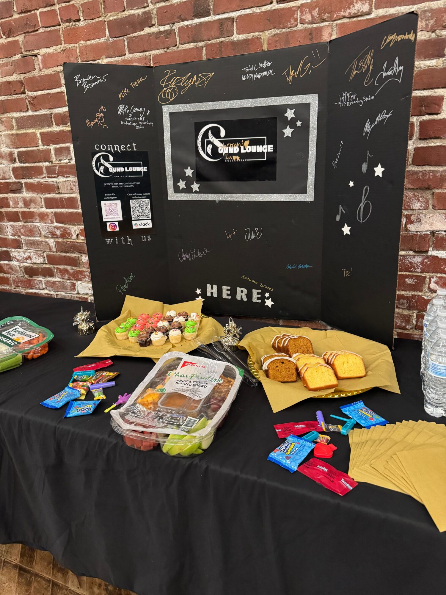 Display table with snacks and a blackboard with handwritten messages and drawings, against a brick wall background.
