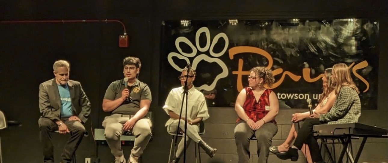 Five people sit on a stage with a black background and a logo, engaged in a discussion or panel.