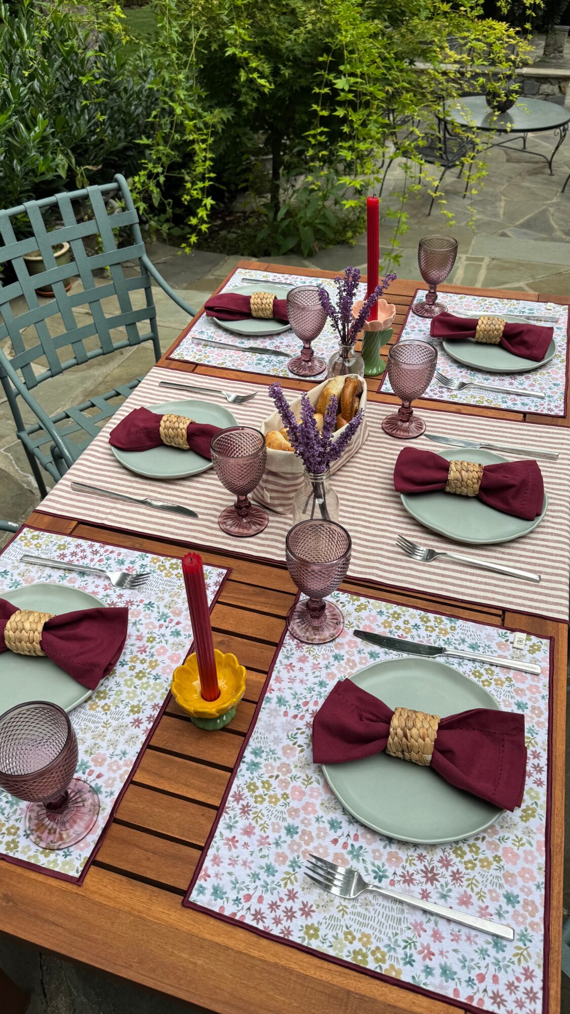 Outdoor dining table set with six place settings, candles, and decorative items, surrounded by greenery and outdoor furniture.