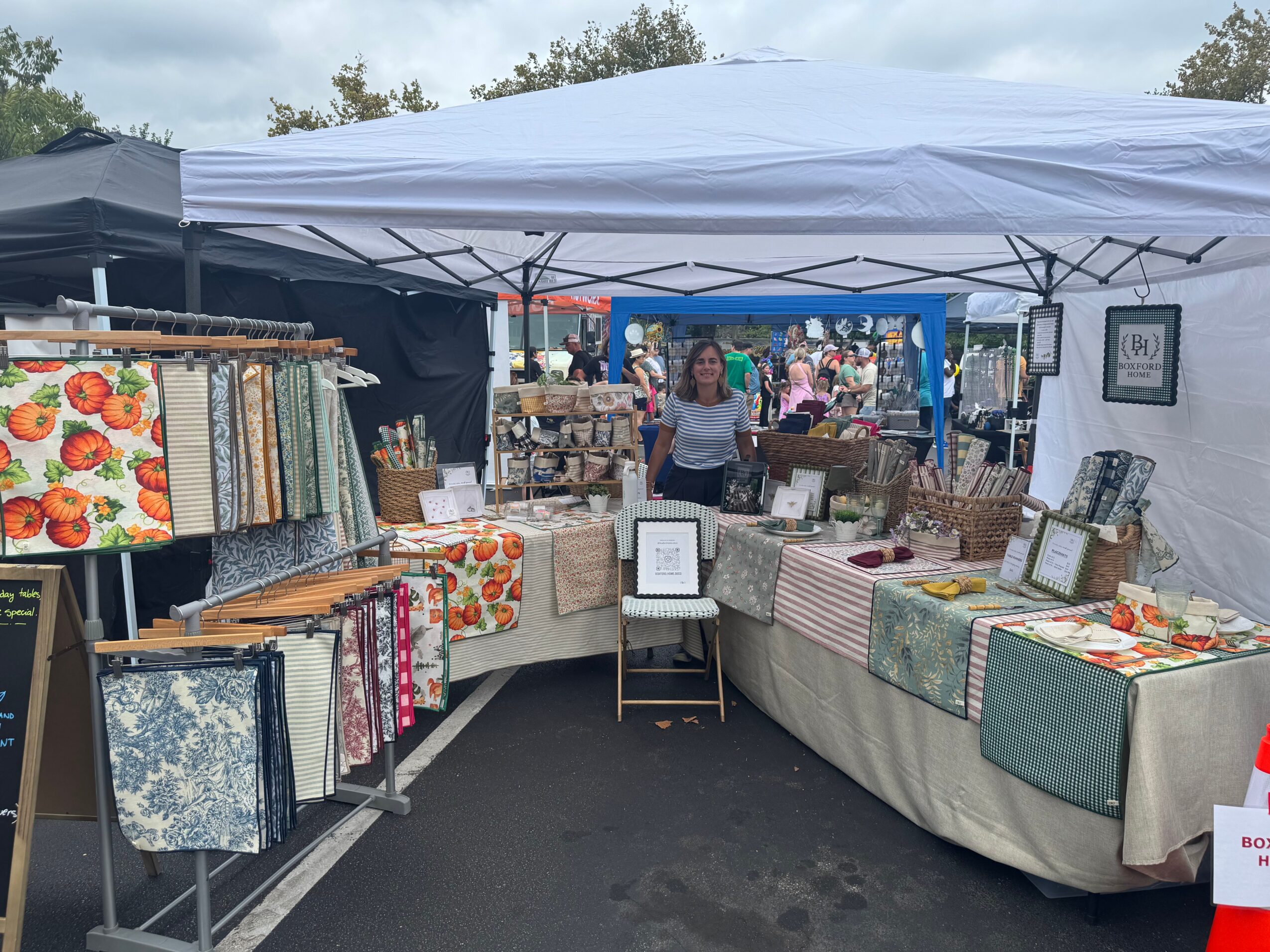 Outdoor market stall with fabric and home decor items under a white canopy, with people and other stalls in background.