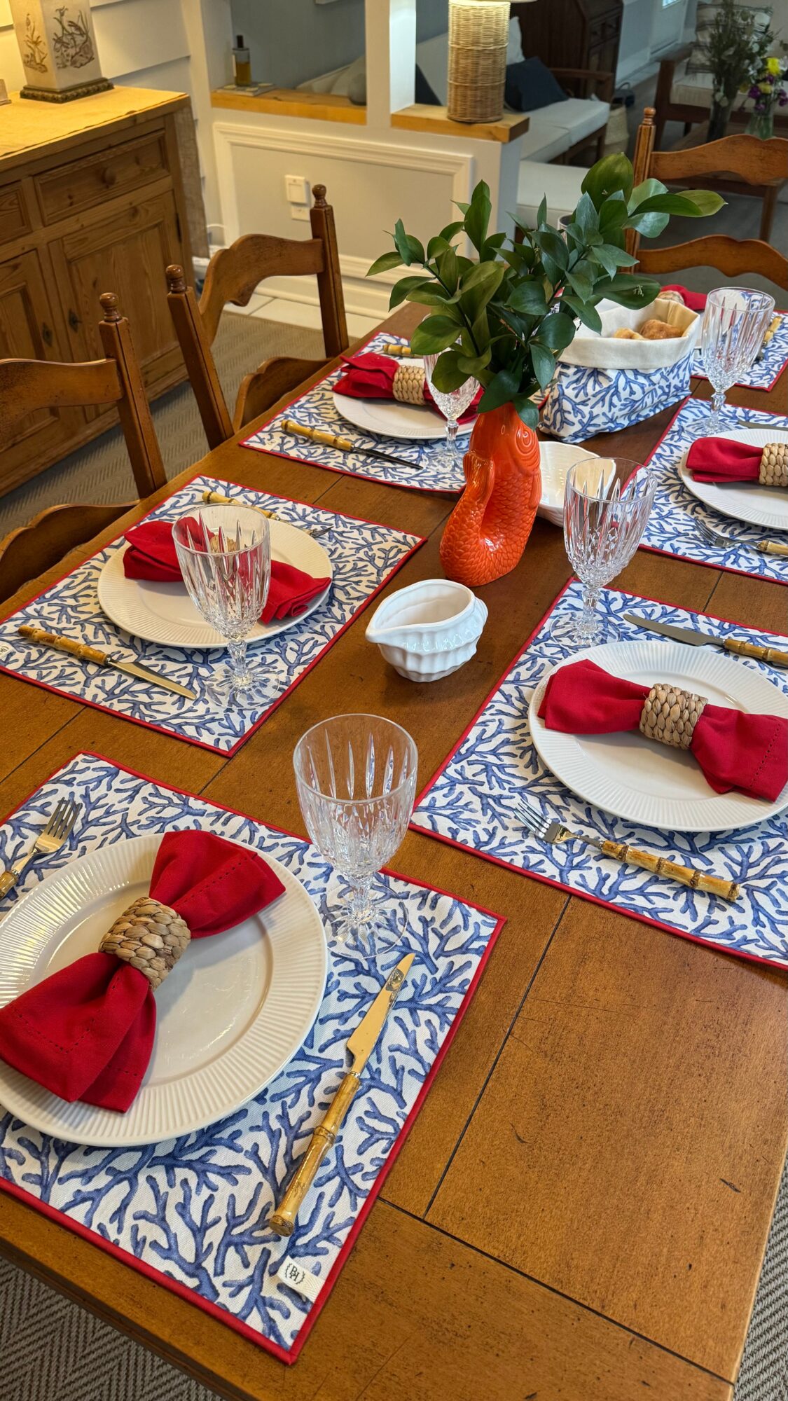 Dining table set with plates, red napkins, gold napkin rings, glasses, a vase with green leaves, and decorative placemats.