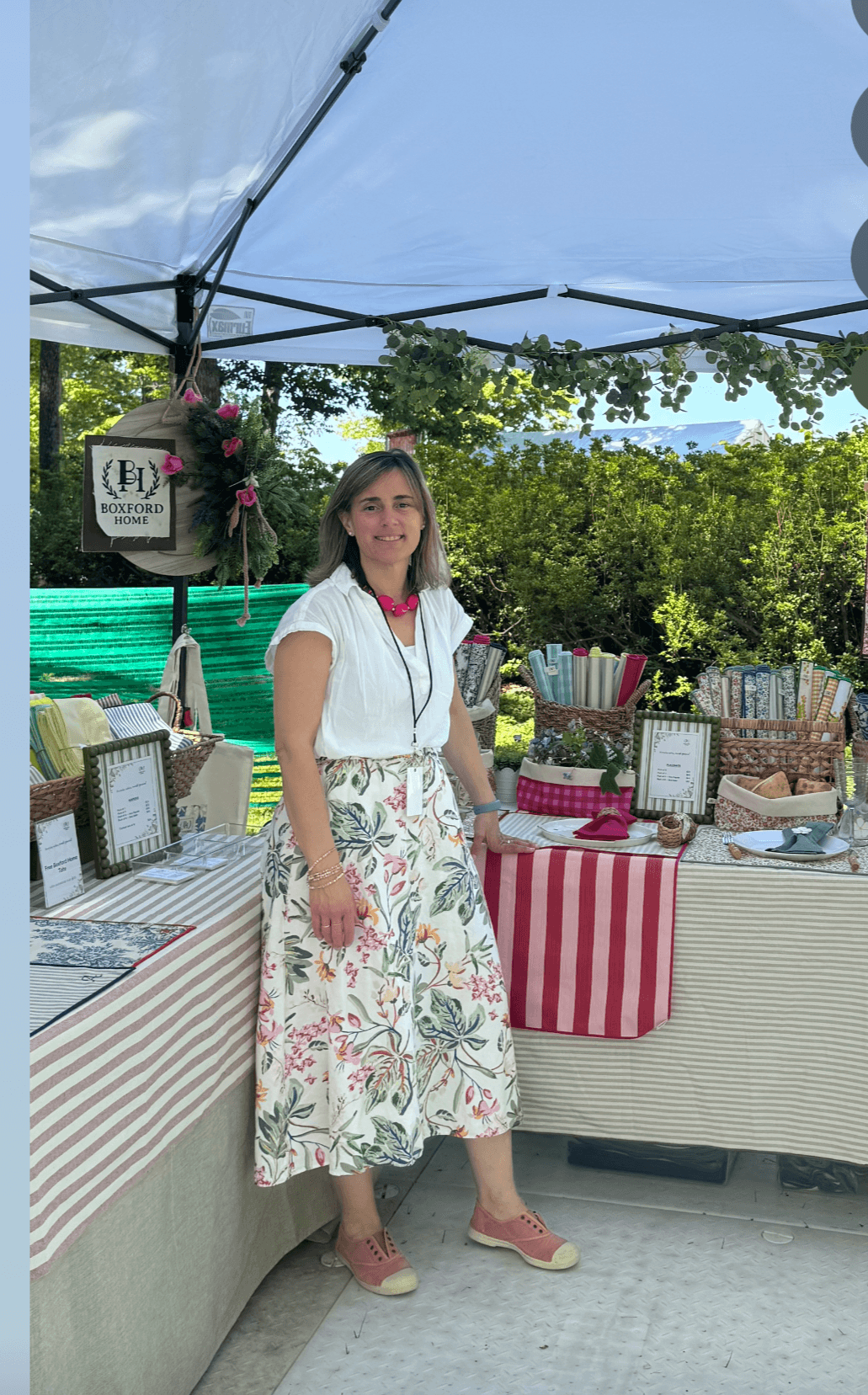Woman standing at outdoor market stall with striped tablecloth, surrounded by framed items and colorful objects, under a canopy with trees in background.