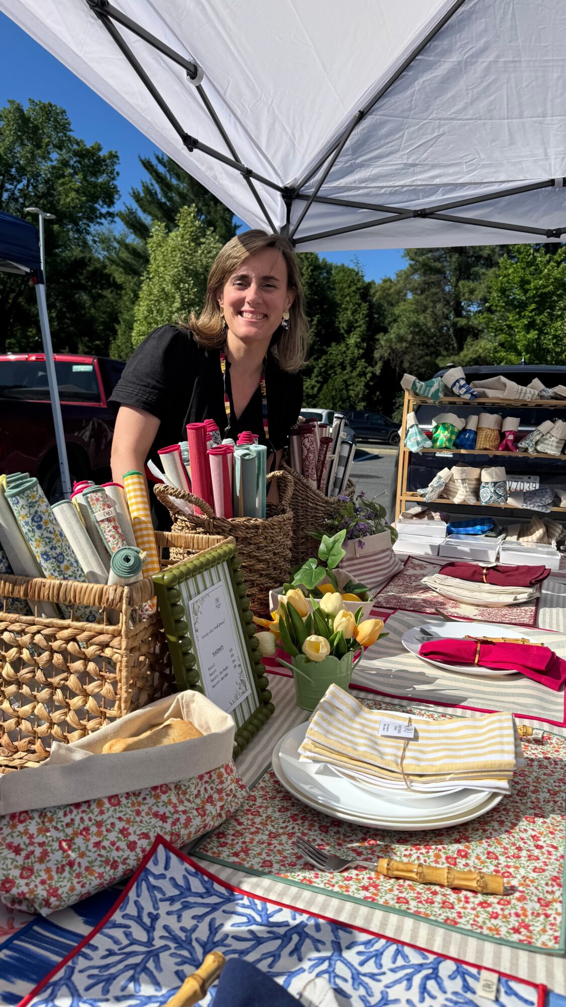 Woman smiling at outdoor market stall with books, flowers, and tableware under a white canopy.