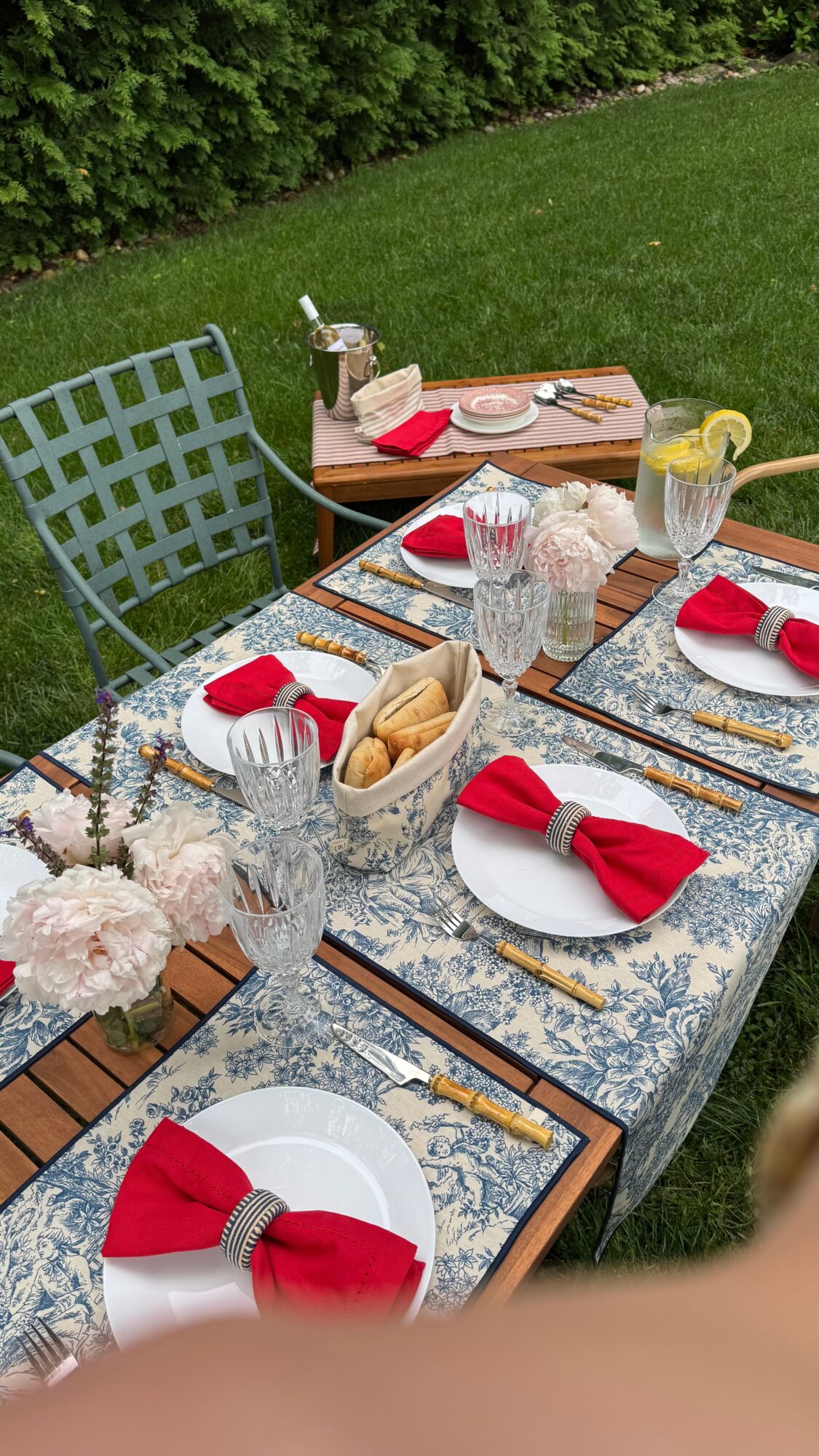 Outdoor table set for a meal with plates, red napkins, glasses, and a pitcher, on a garden patio with grass and trees.