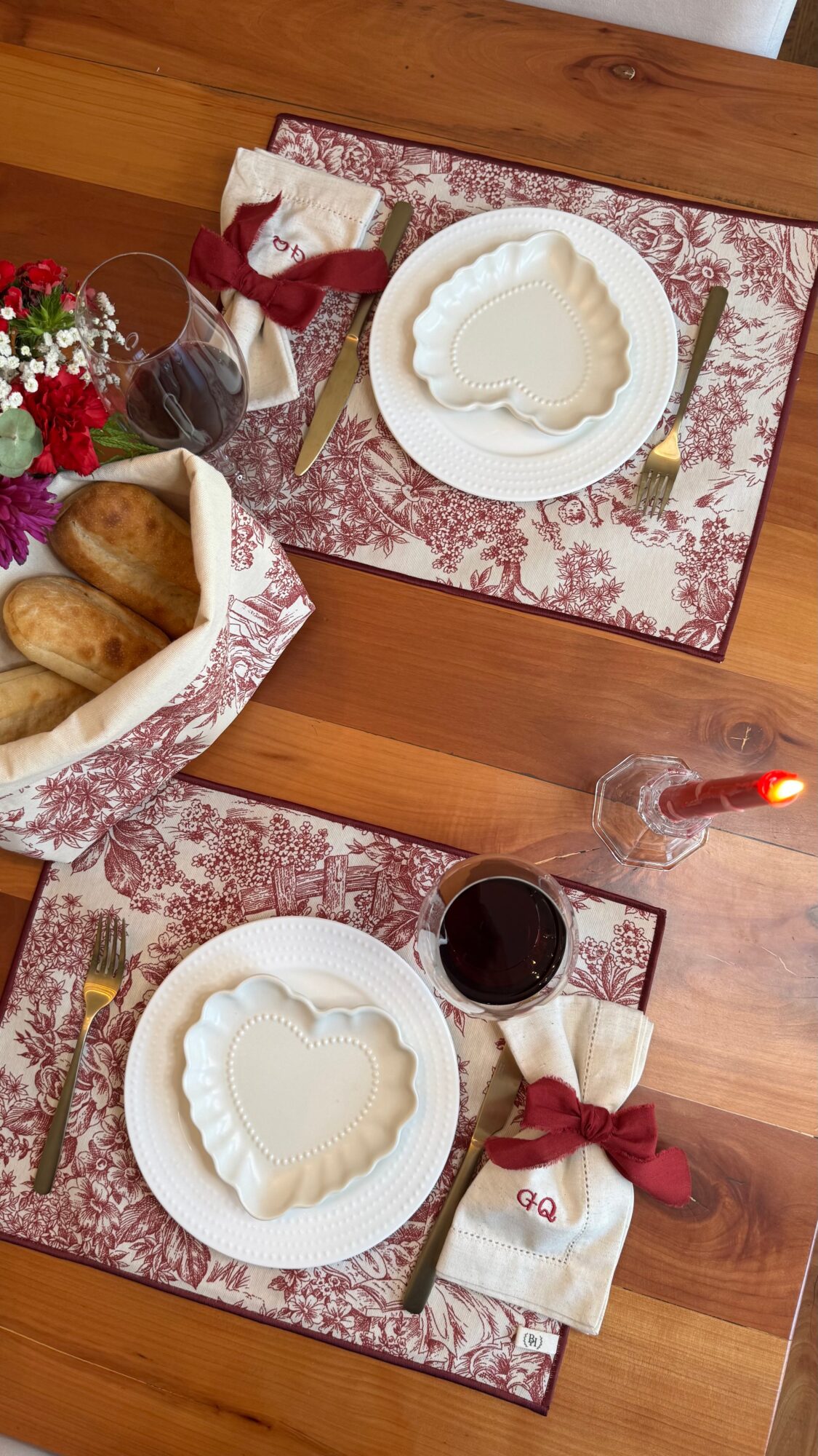Table setting with two plates, heart-shaped desserts, wine, bread basket, flowers, and candles on a wooden table.