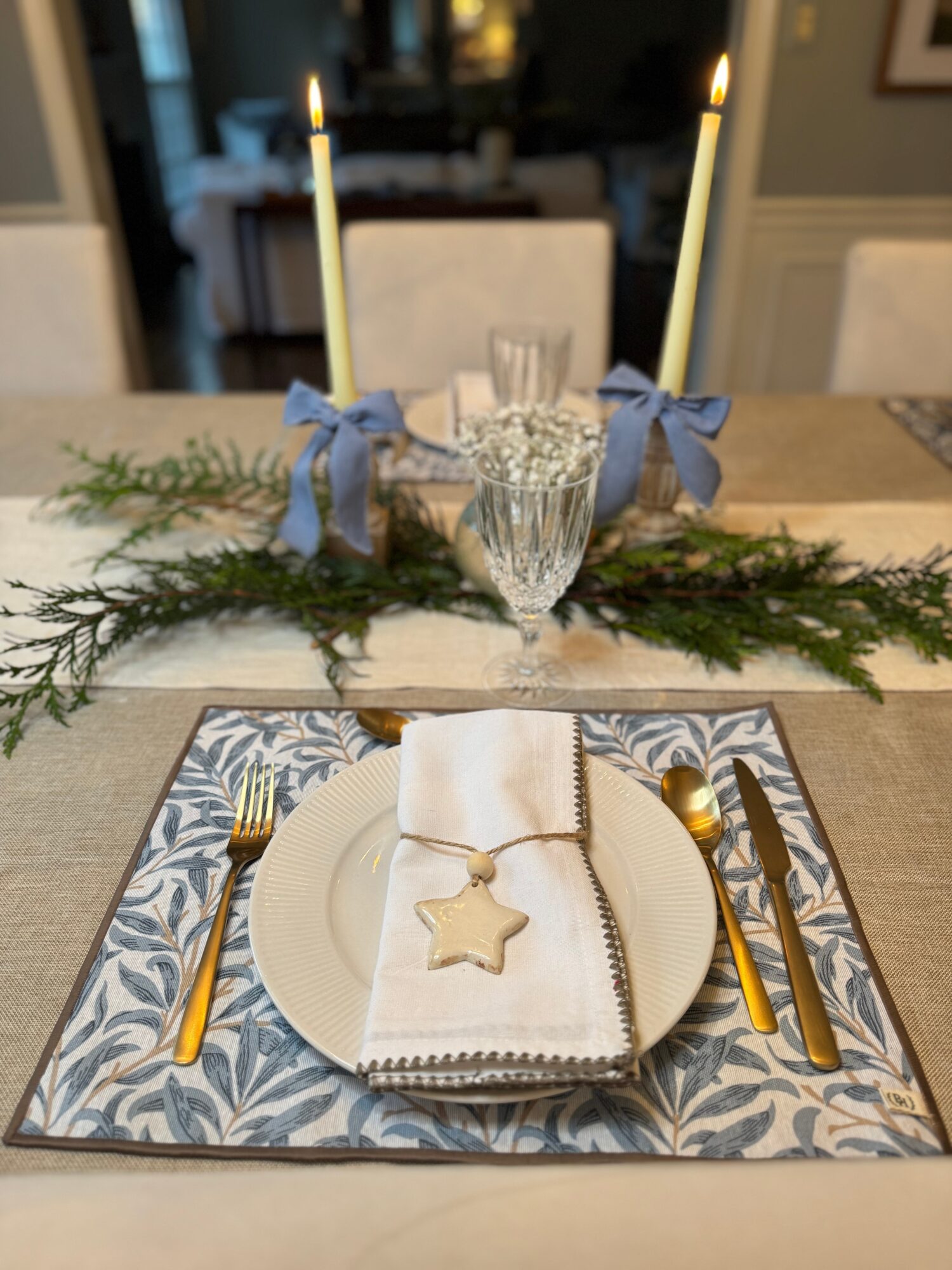 Festive table setting with candles, greenery, a glass, and a decorated plate with a star ornament, gold cutlery, and a patterned placemat.