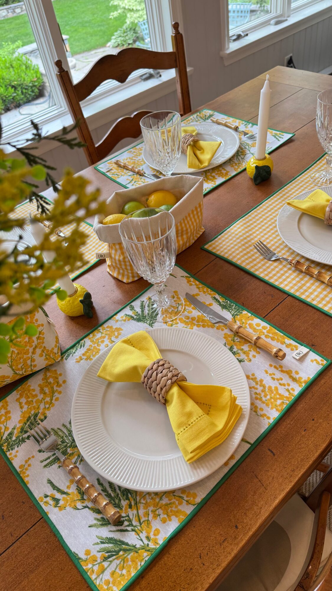 Dining table set with plates, yellow napkins, glasses, and decorative items, near windows with greenery outside.