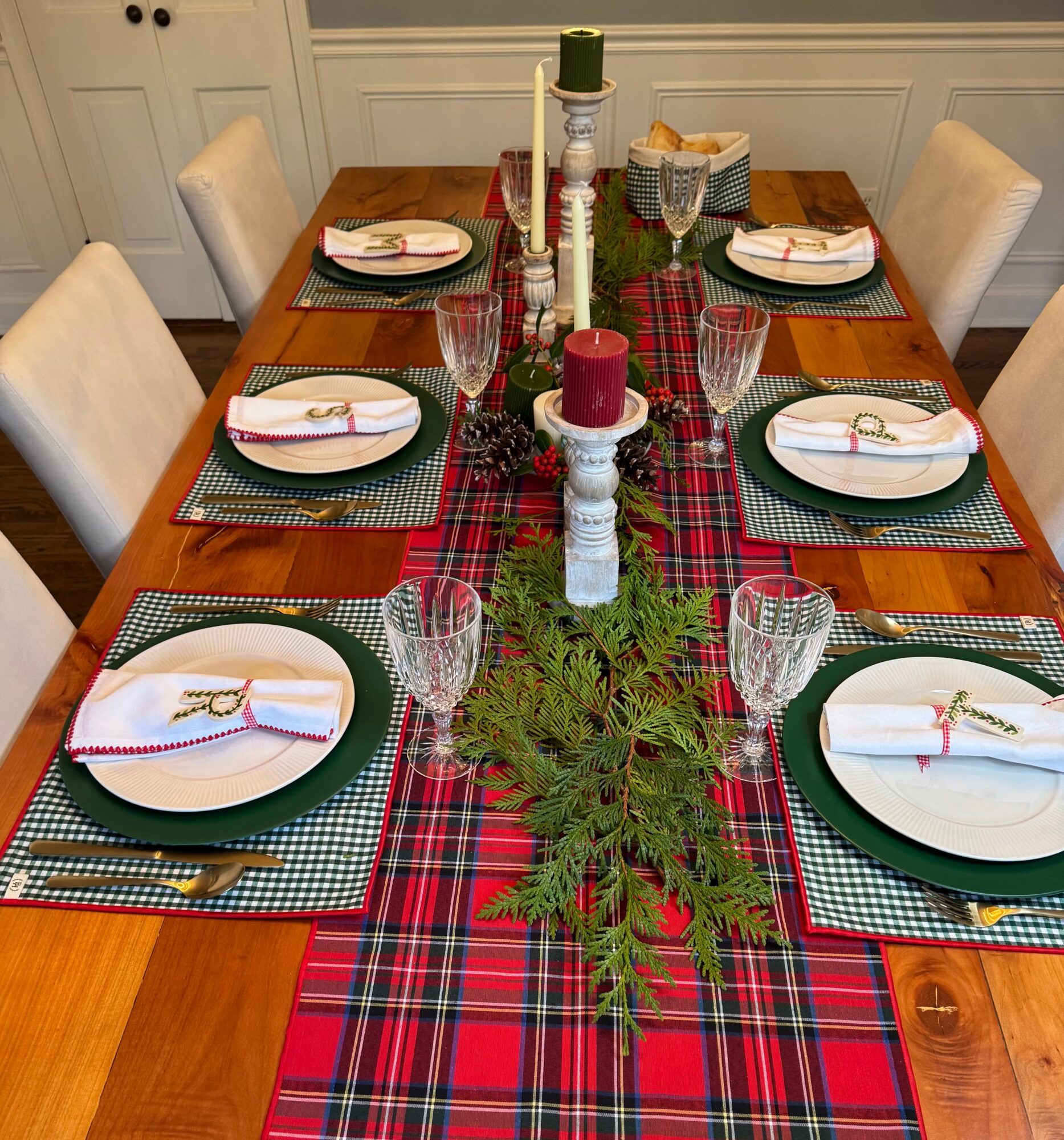 Dining table set with six place settings, a red and green plaid table runner, candles, and greenery centerpiece.