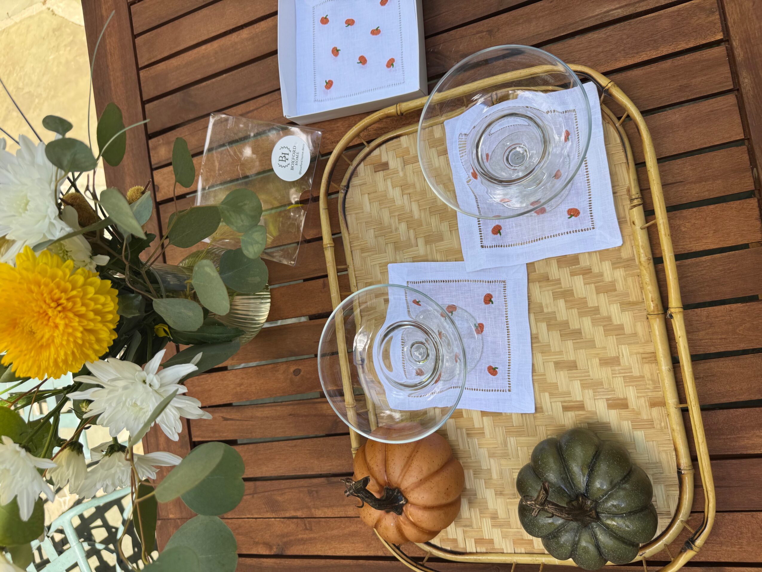 Table with a tray, two glasses, a flower arrangement, and two pumpkins on a wooden surface.