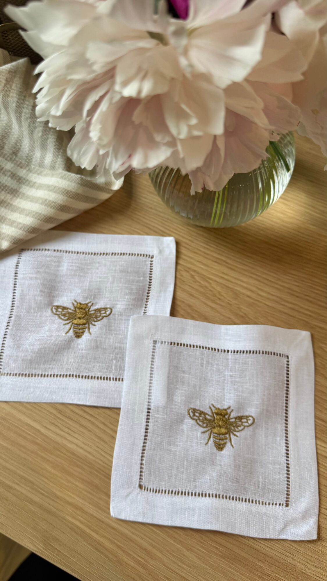 Two white cloth napkins with embroidered gold bees, placed on a wooden table near a vase of pink flowers.