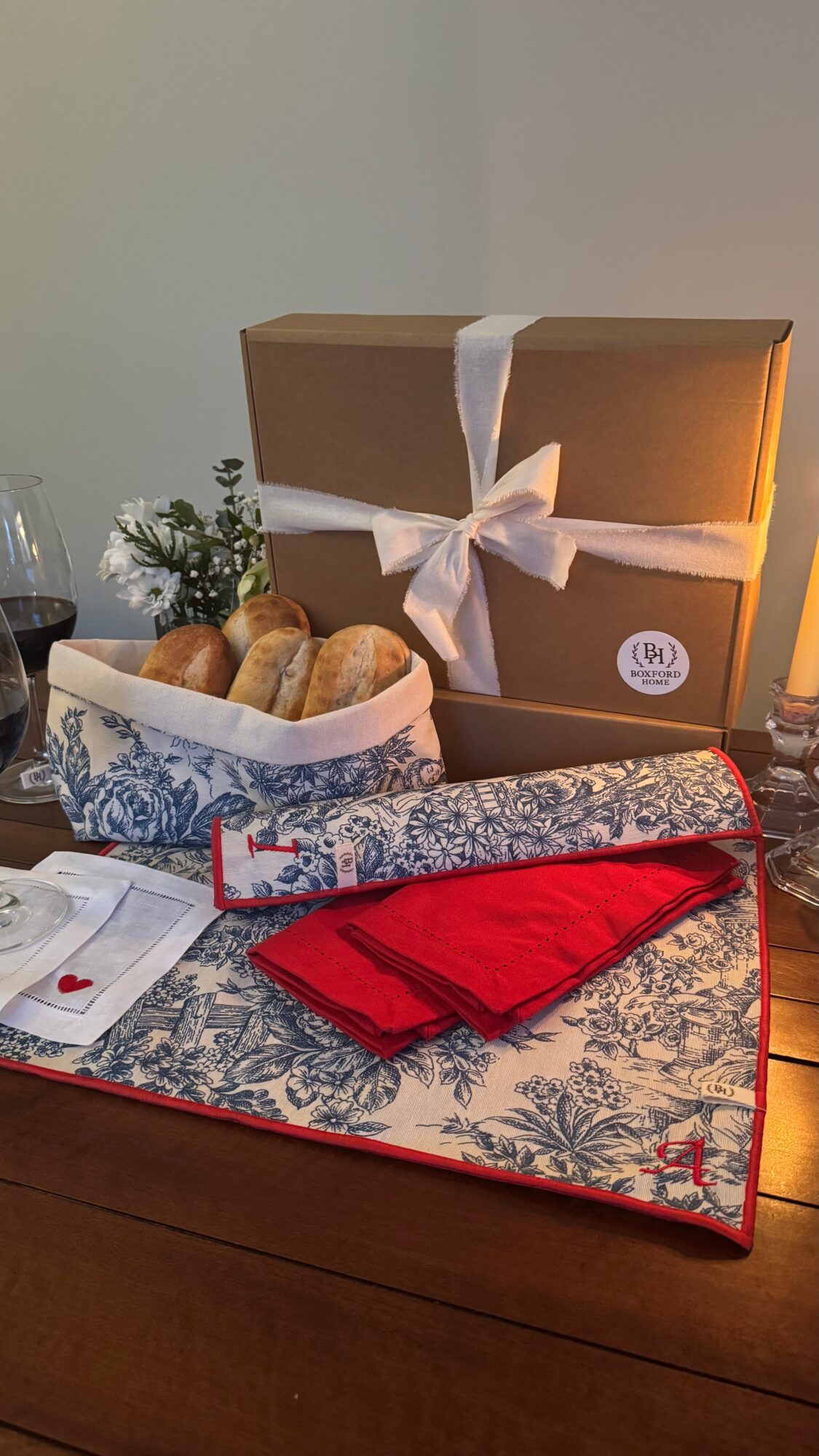 Gift box with white ribbon, bread basket, red cloth, and decorative table runner on wooden table.