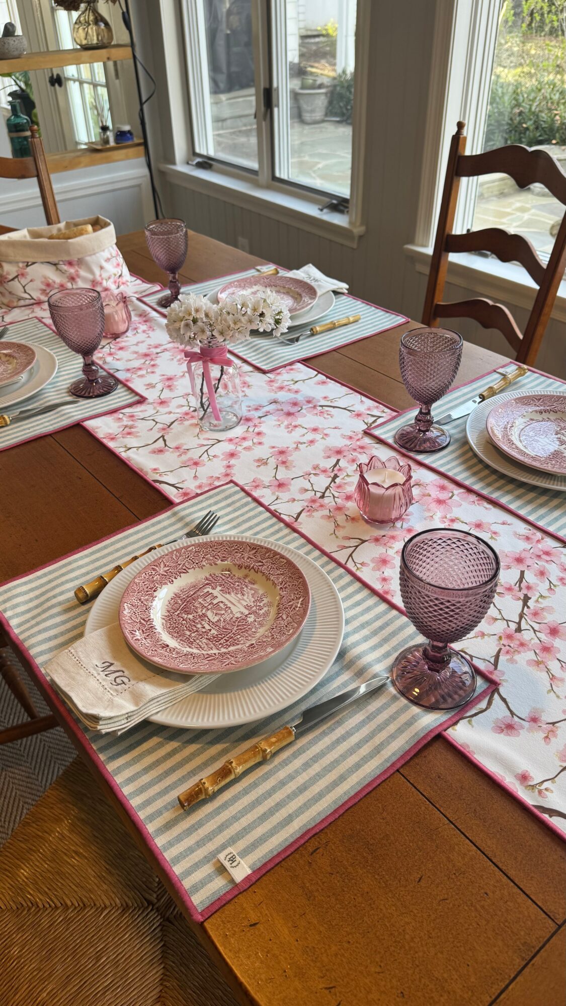 Dining table set with pink and purple glassware, plates, and a floral table runner, near a window with sunlight.