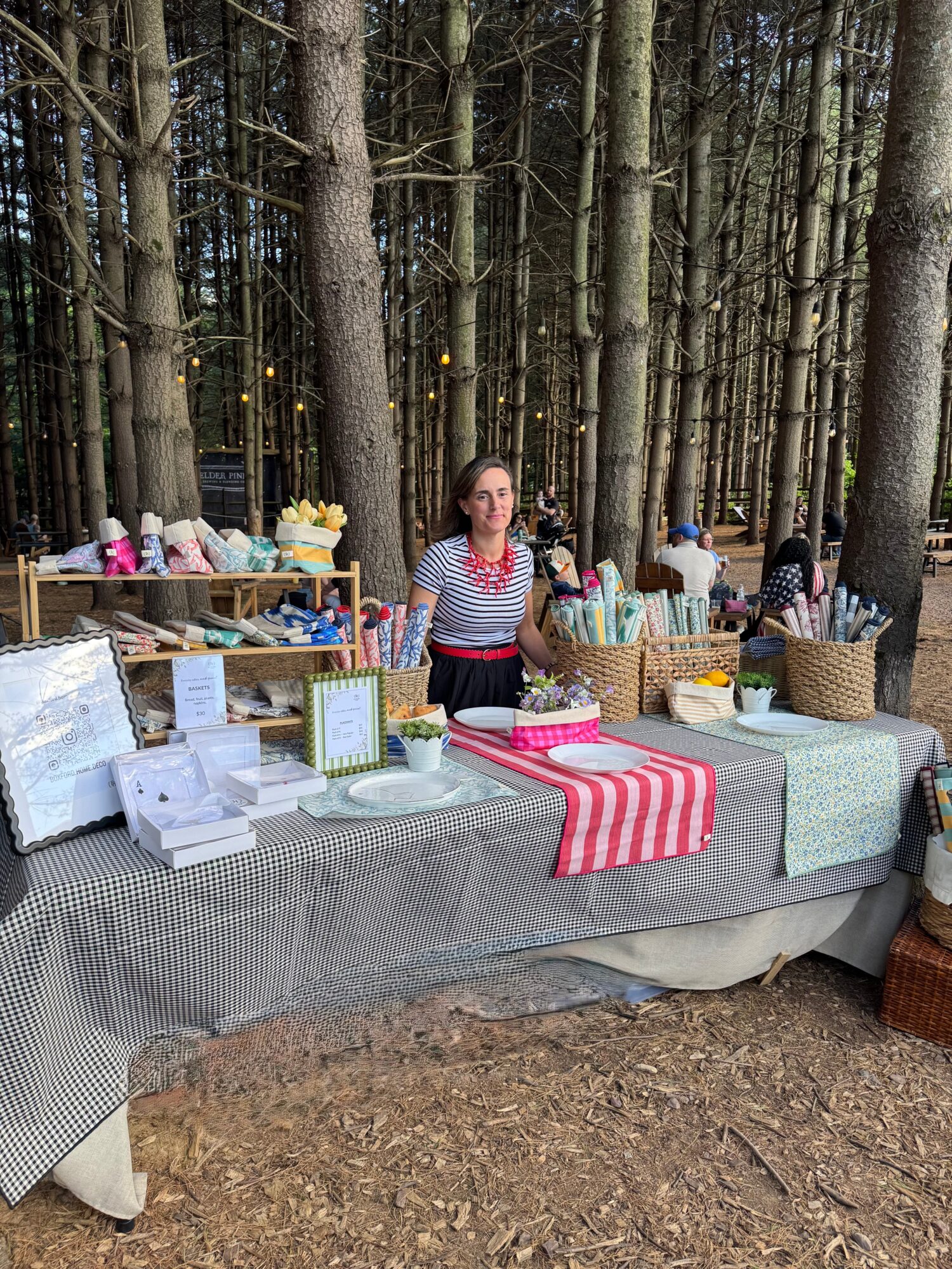 Woman standing behind a table with baked goods and baskets in a forest setting, trees in the background.