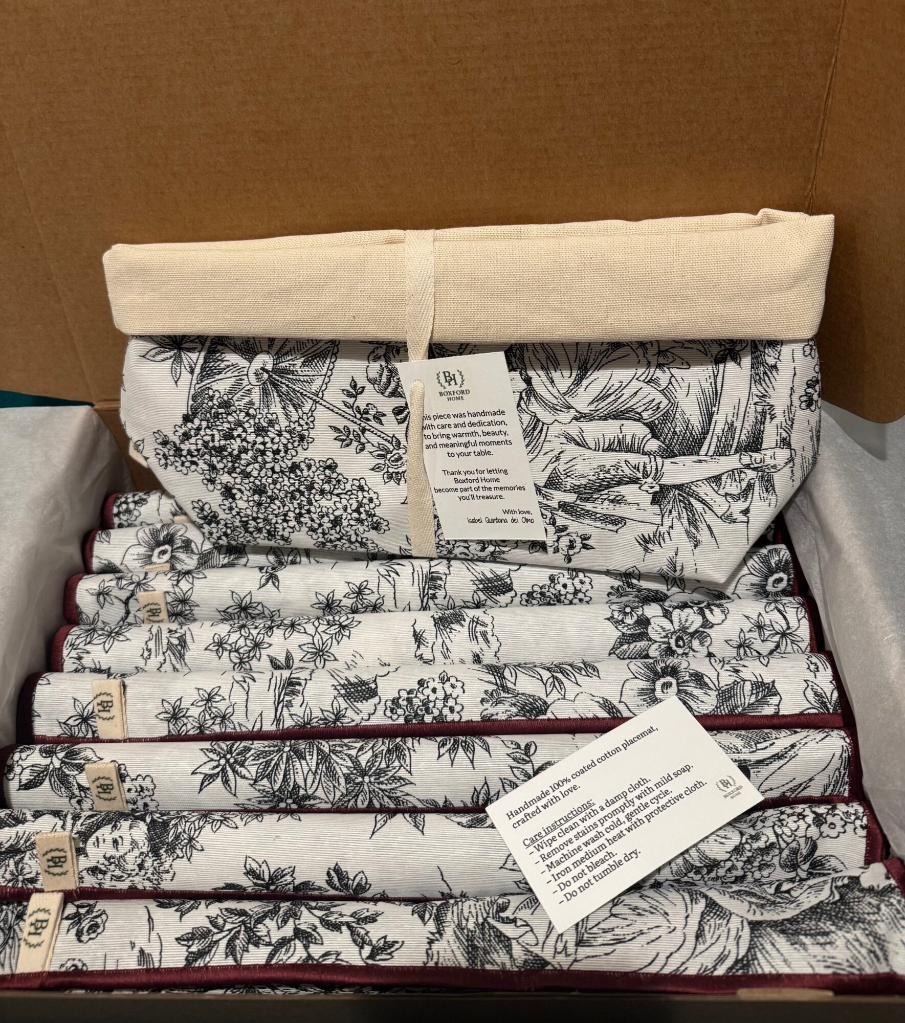 Stack of folded fabric or linens with floral patterns, placed inside a box, with tags attached.
