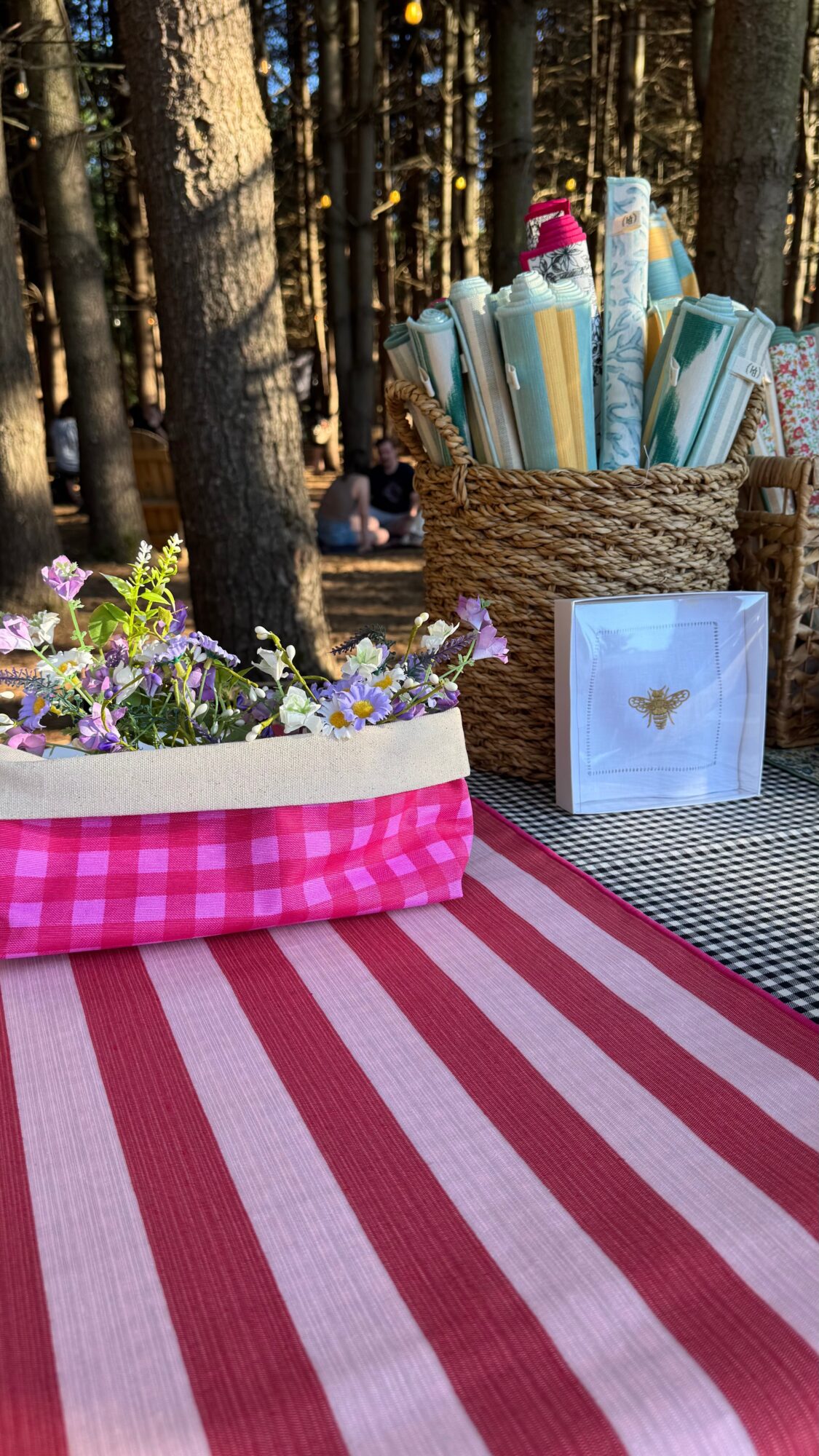 Picnic setup with a striped red and white blanket, flower pot, and basket of rolled papers in a forest.