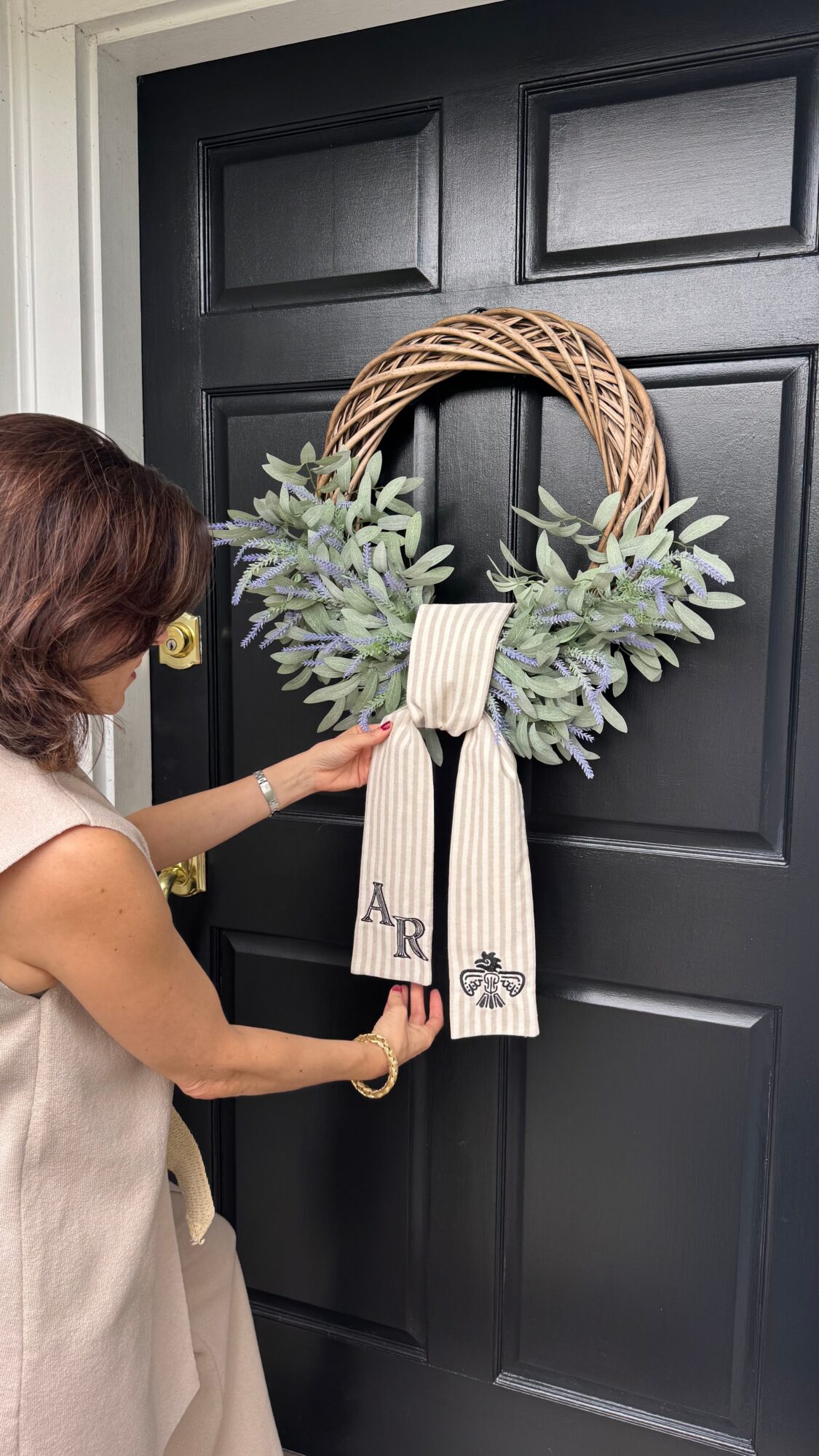 Person hanging a decorative wreath with greenery and a ribbon on a black door.