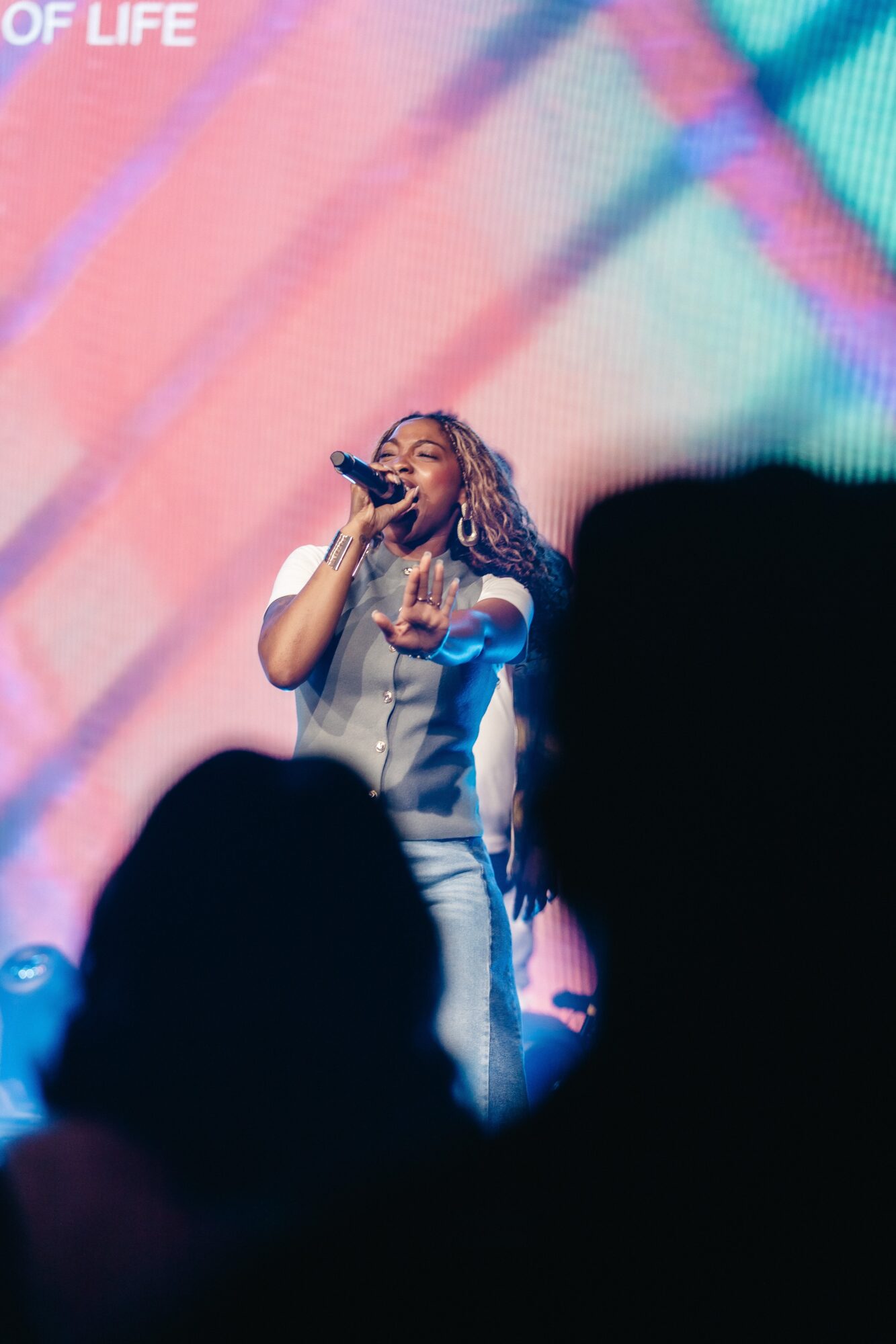 A woman with curly hair sings into a microphone on stage, with colorful lights in the background.