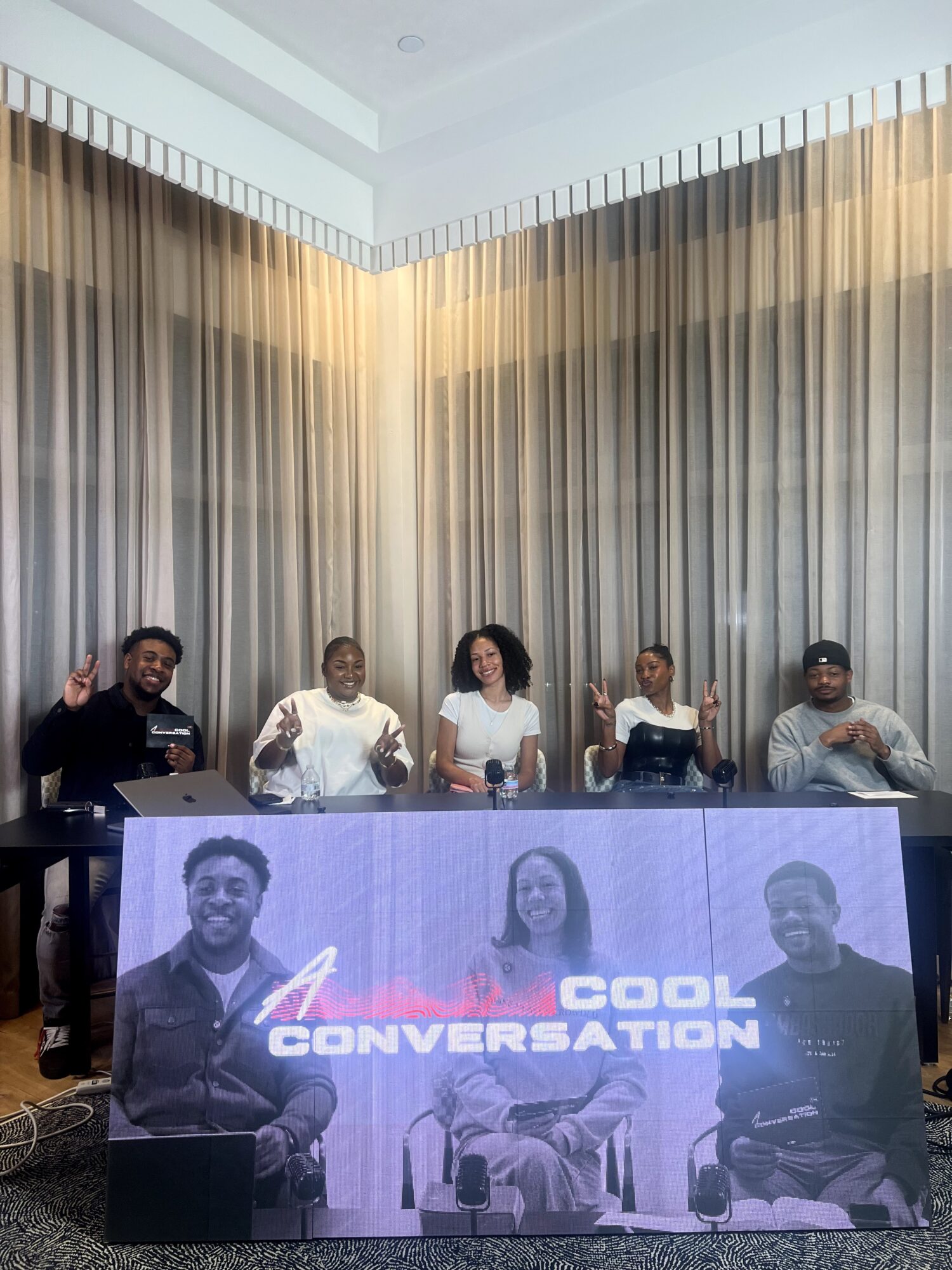 Five people sitting behind a table with a large banner displaying two men and the words 'Cool Conversation'.
