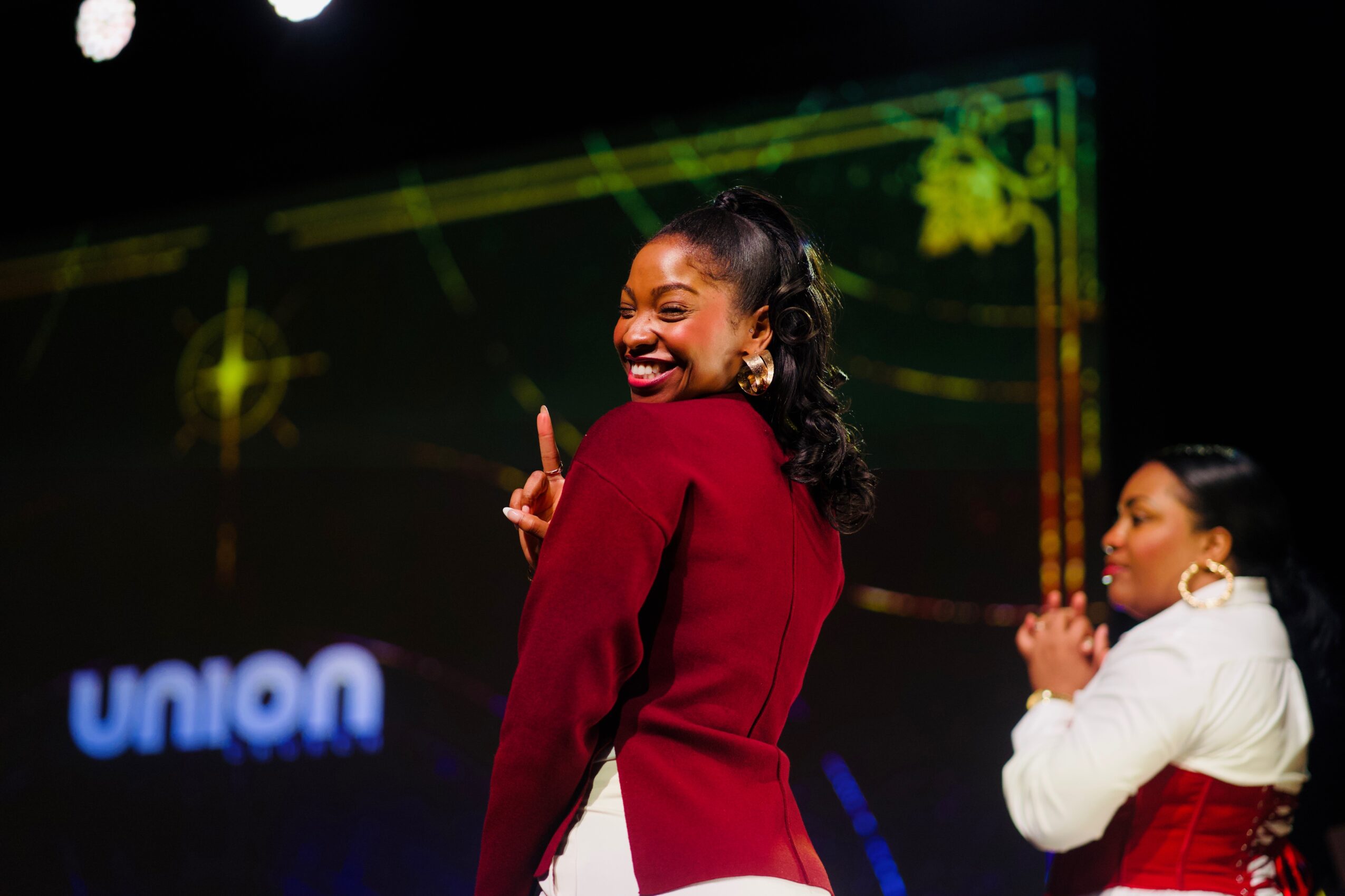 Two women smiling and posing on stage, one in a red blazer and the other in a white top, with a dark background and stage lights.