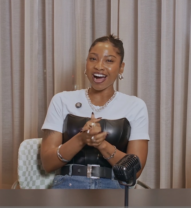 Smiling woman with jewelry, sitting at a table, in front of curtains, wearing a white top and black leather bustier.