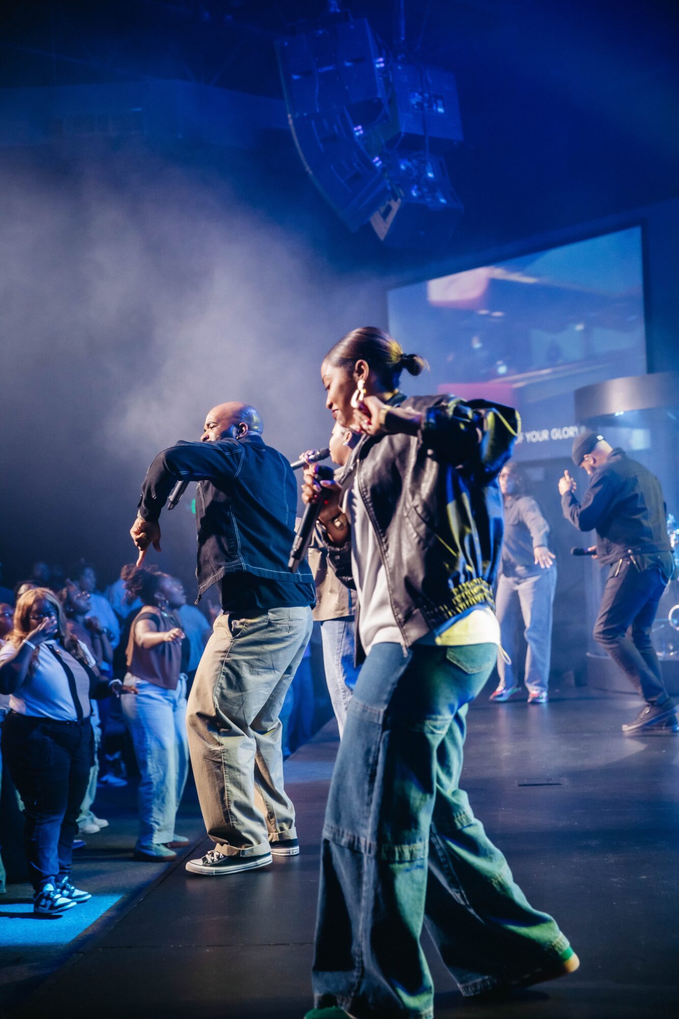 People dancing on stage with colorful lights and a large screen in the background, fog effects present.